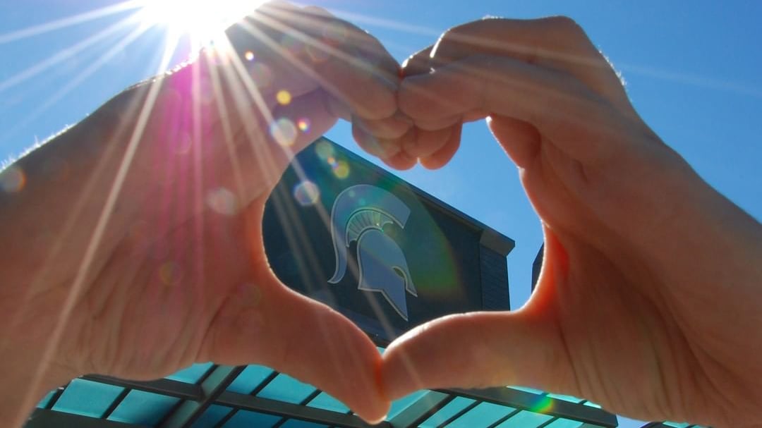 A person's hands formed in the shape of a heart in front of Spartan stadium