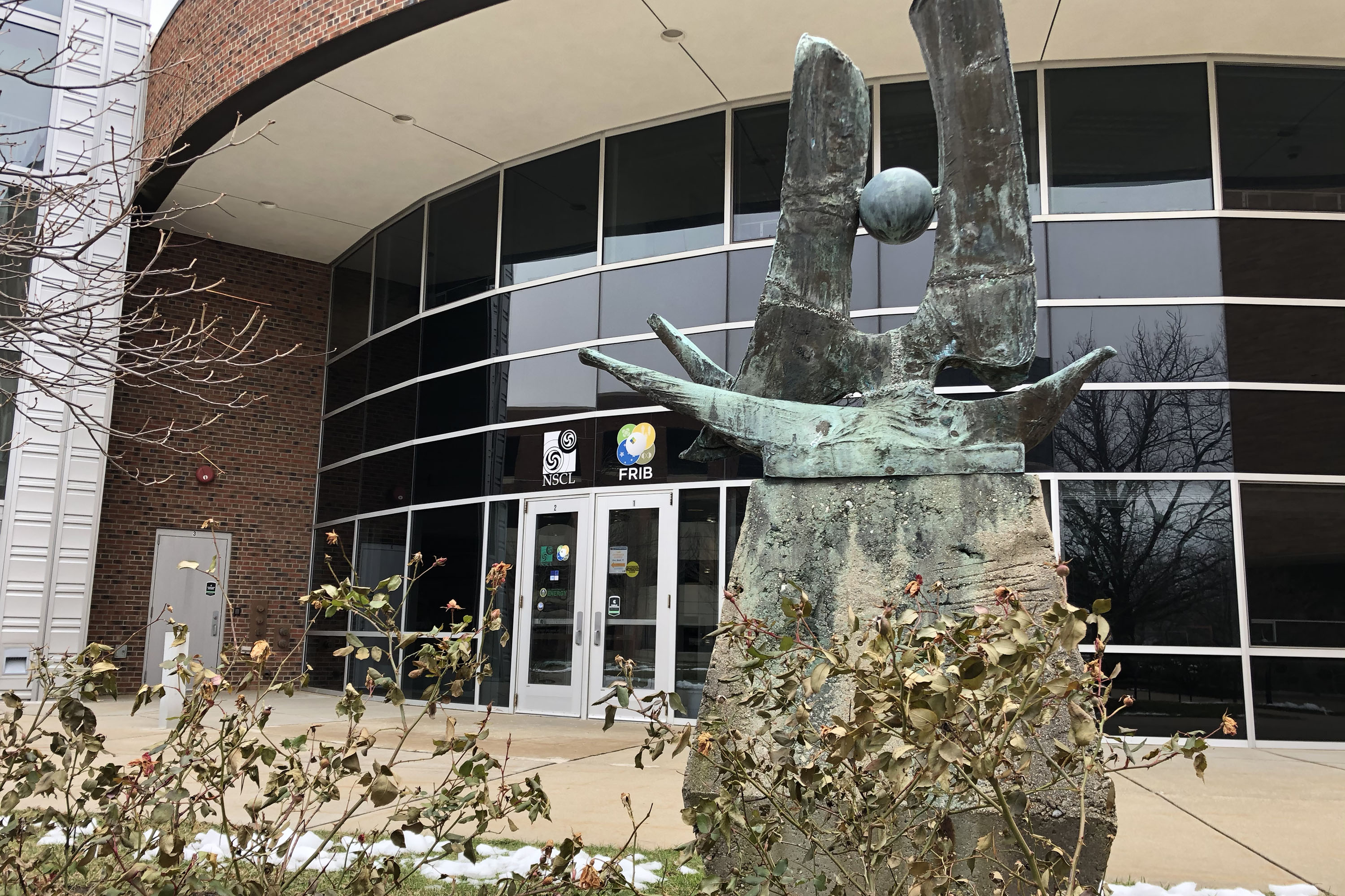 Modern shape sculpture of a ball between curved pillars outside the entrance to the NSCL and FRIB.