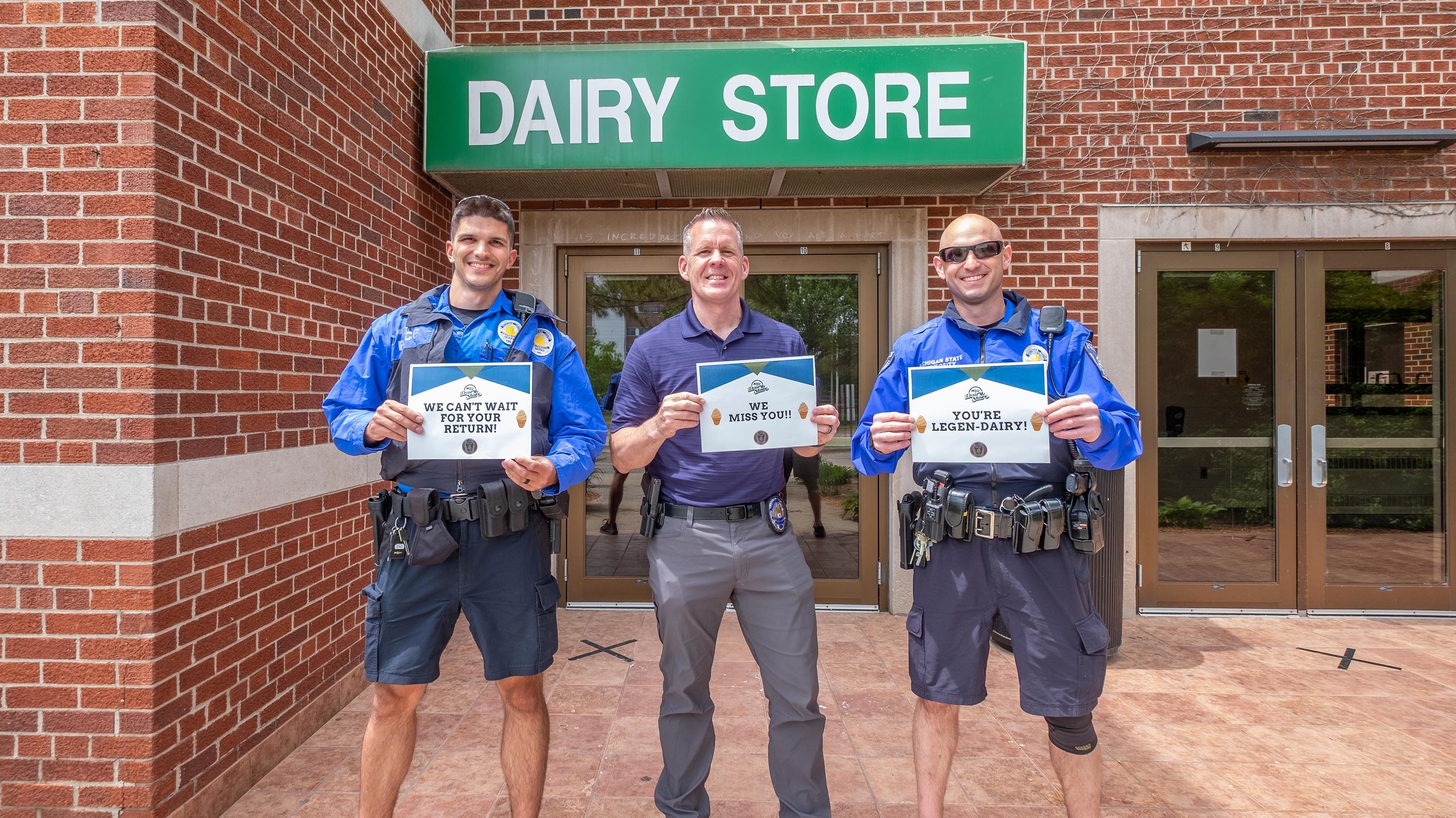 MSU campus police holding signs in front of the Dairy Store. The signs read 'We can't wait for your return', 'we miss you', and 'you're legen-dairy'..