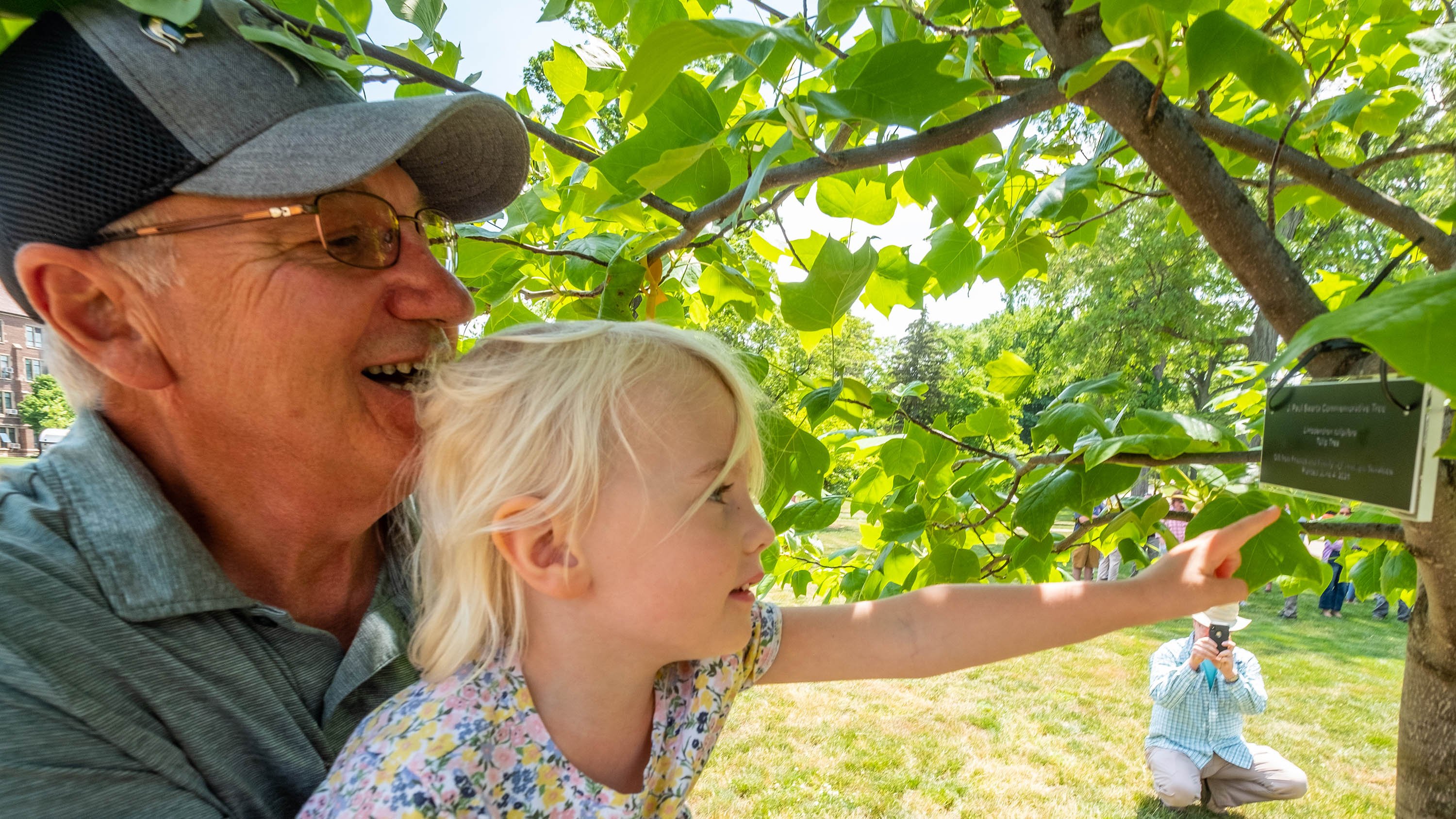 Paul Swartz holding up his granddaughter to examine the plaque on the tree that honors him.