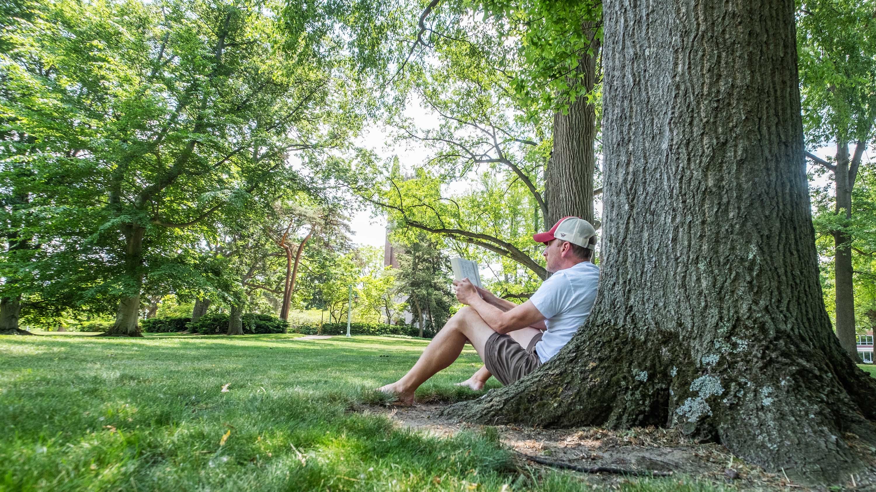 A person reading a book while sitting against a tree on campus, Beaumont Tower in the distance