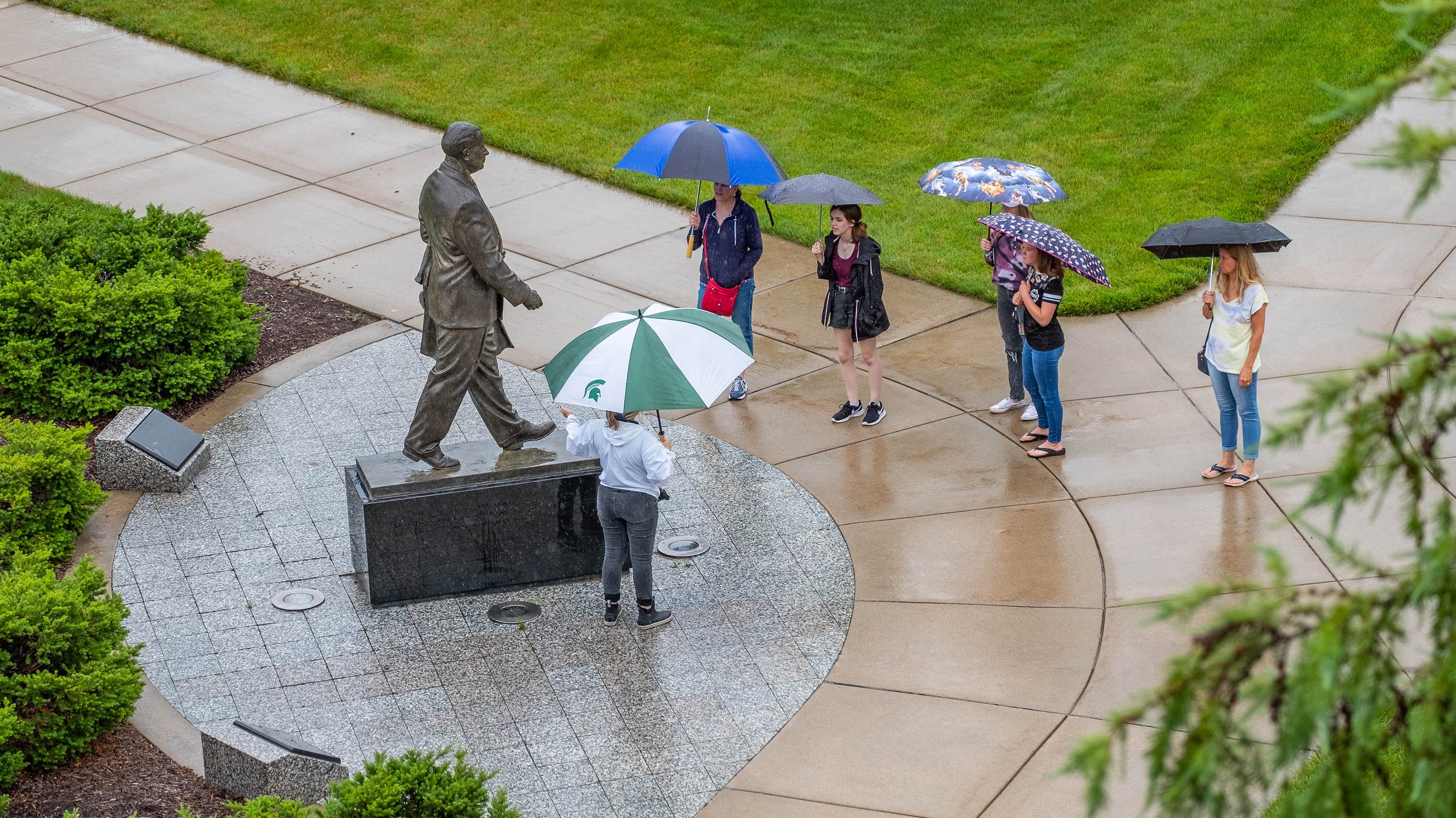 Campus tour guide showing people a statue of John Hannah on a rainy day, under umbrellas