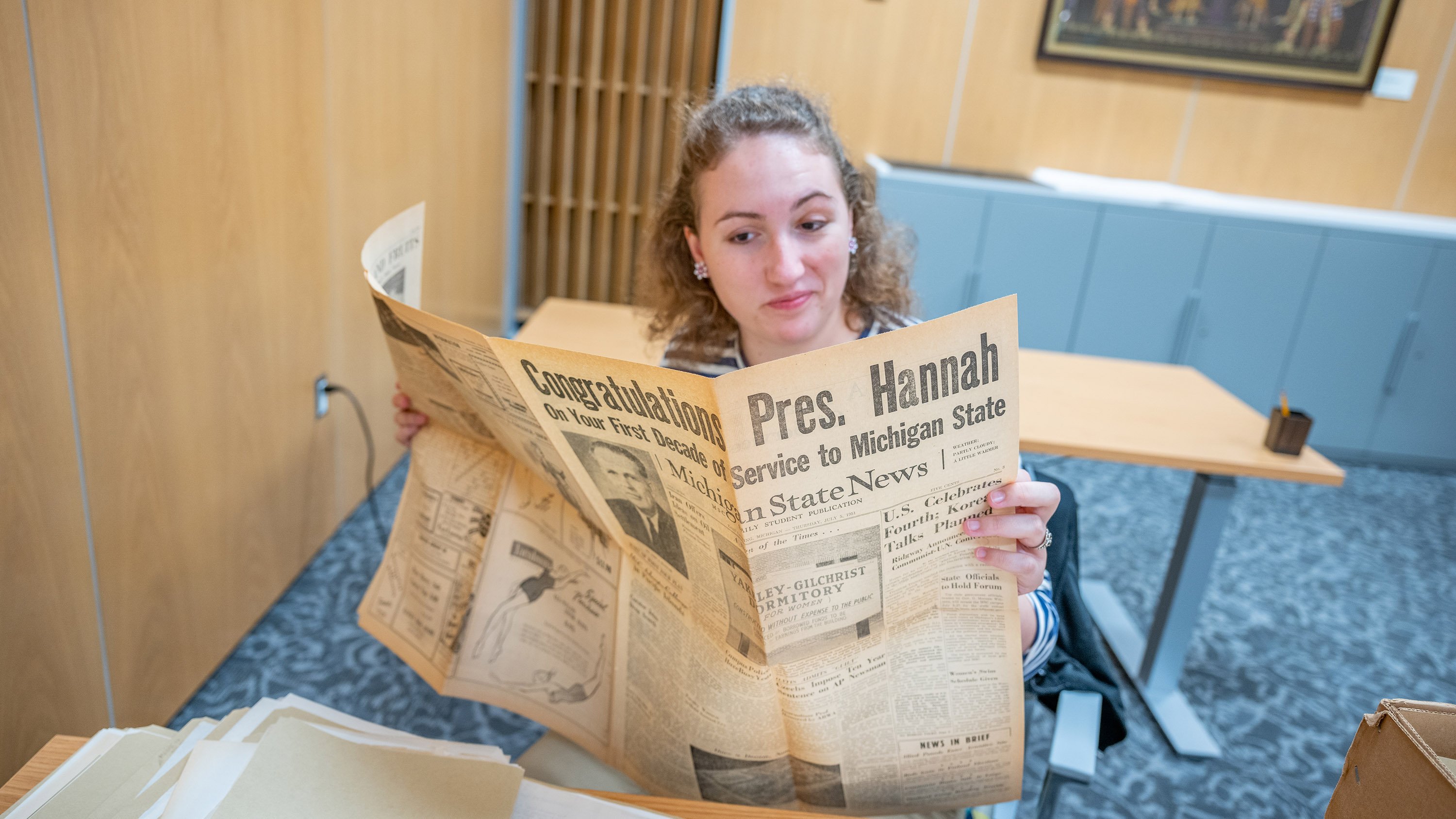 Hannah Zimmerman reading a newspaper with the headline 'Congratulations Pres. Hannah, on your first decade of service to Michigan State'