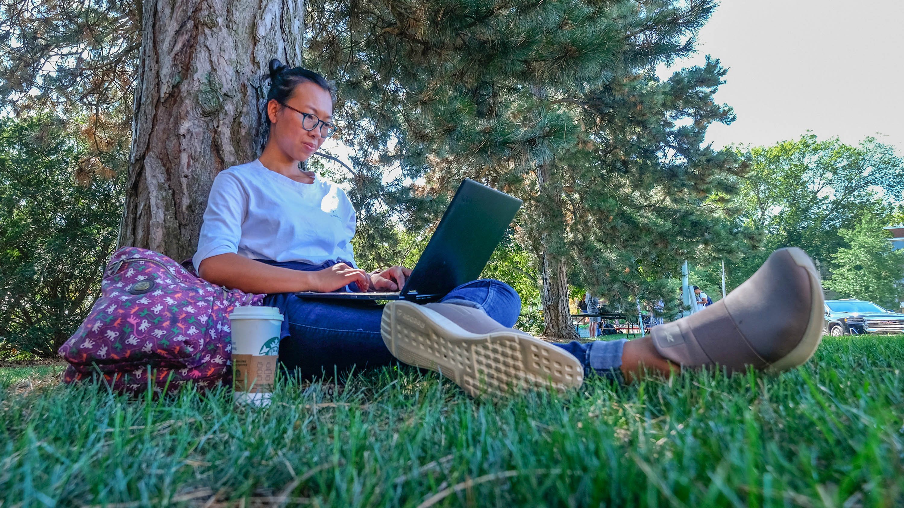 Student sitting against a tree with a laptop and coffee