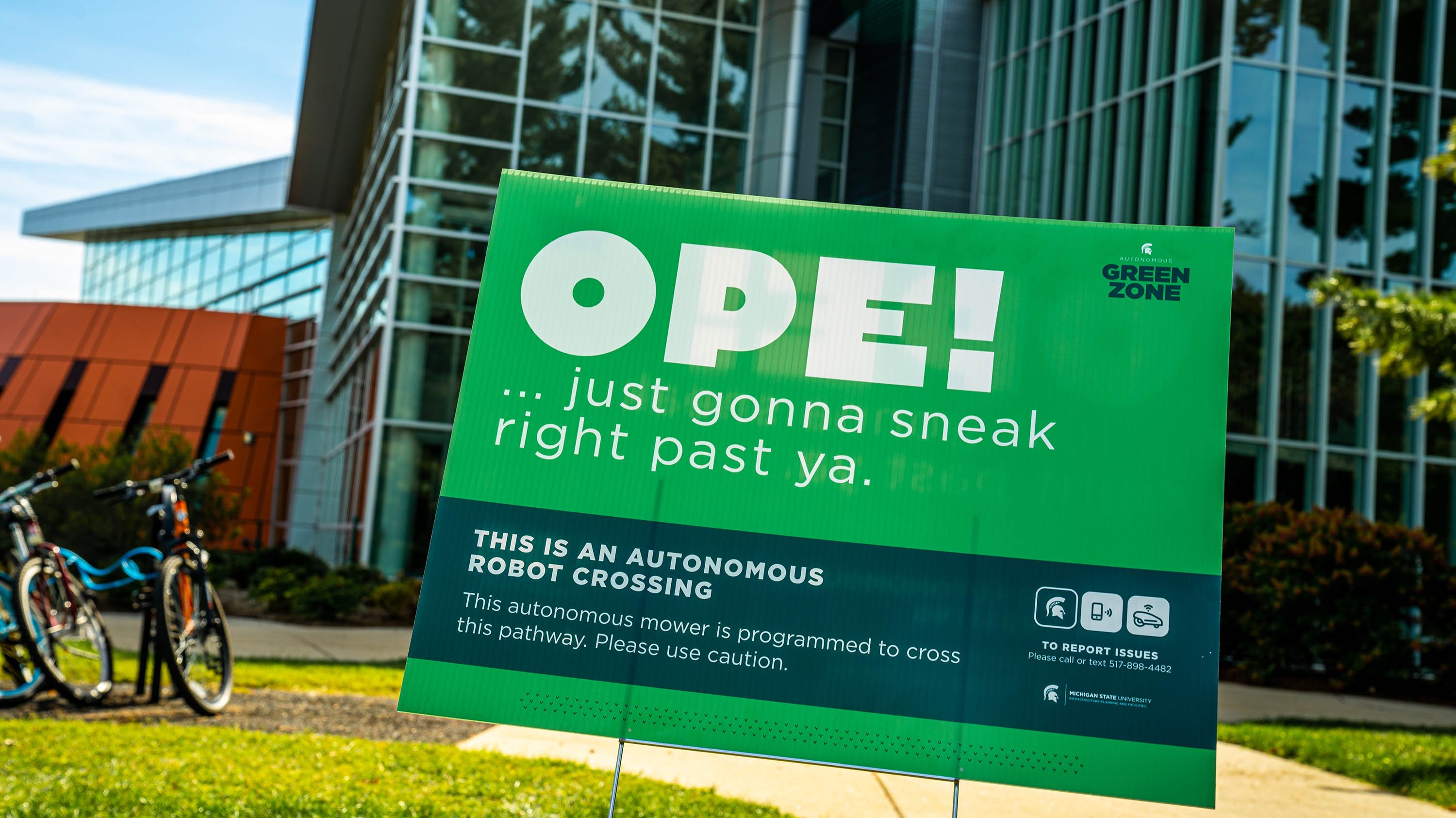 Sign posted in grass with text that reads 'Ope! Just gonna sneak right past ya' with more information about robotic lawnmowers on campus