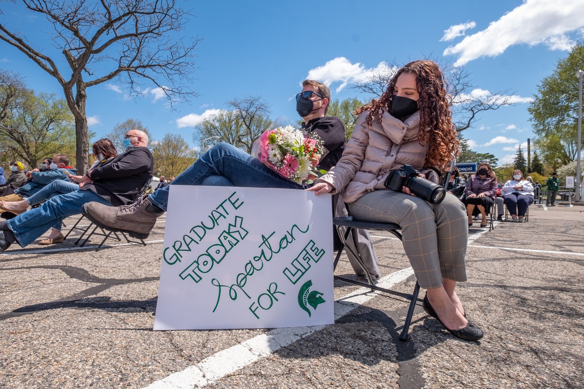 A male sitting in a chair has a bouquet of flowers on his lap. A female sitting next to him as a sign saying ?Graduate today Spartan for life.? 