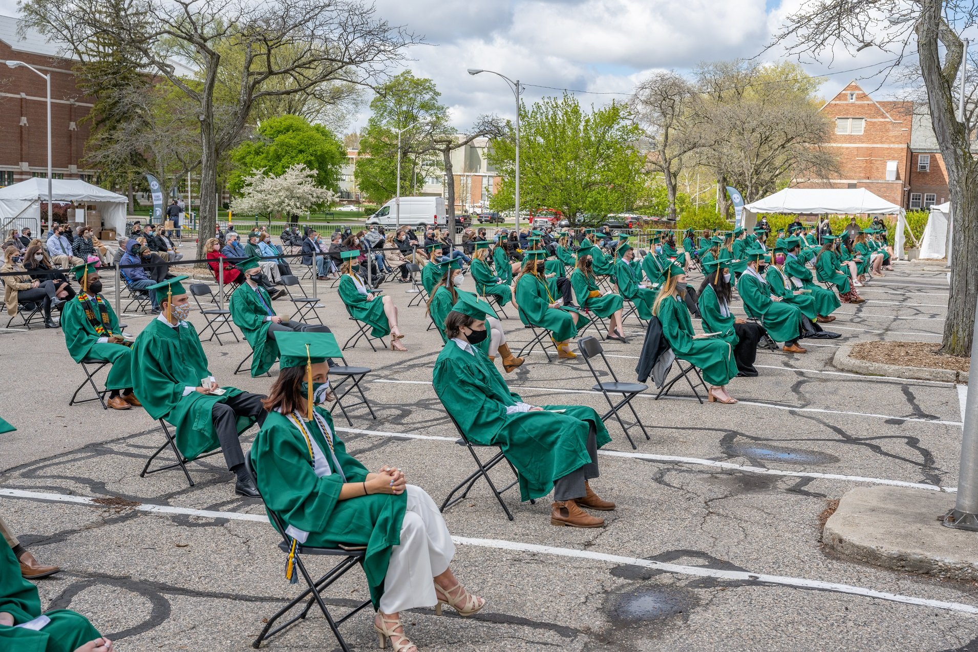 People in green caps and gowns sit spread apart in rows of chairs in the MSU Auditorium parking lot.