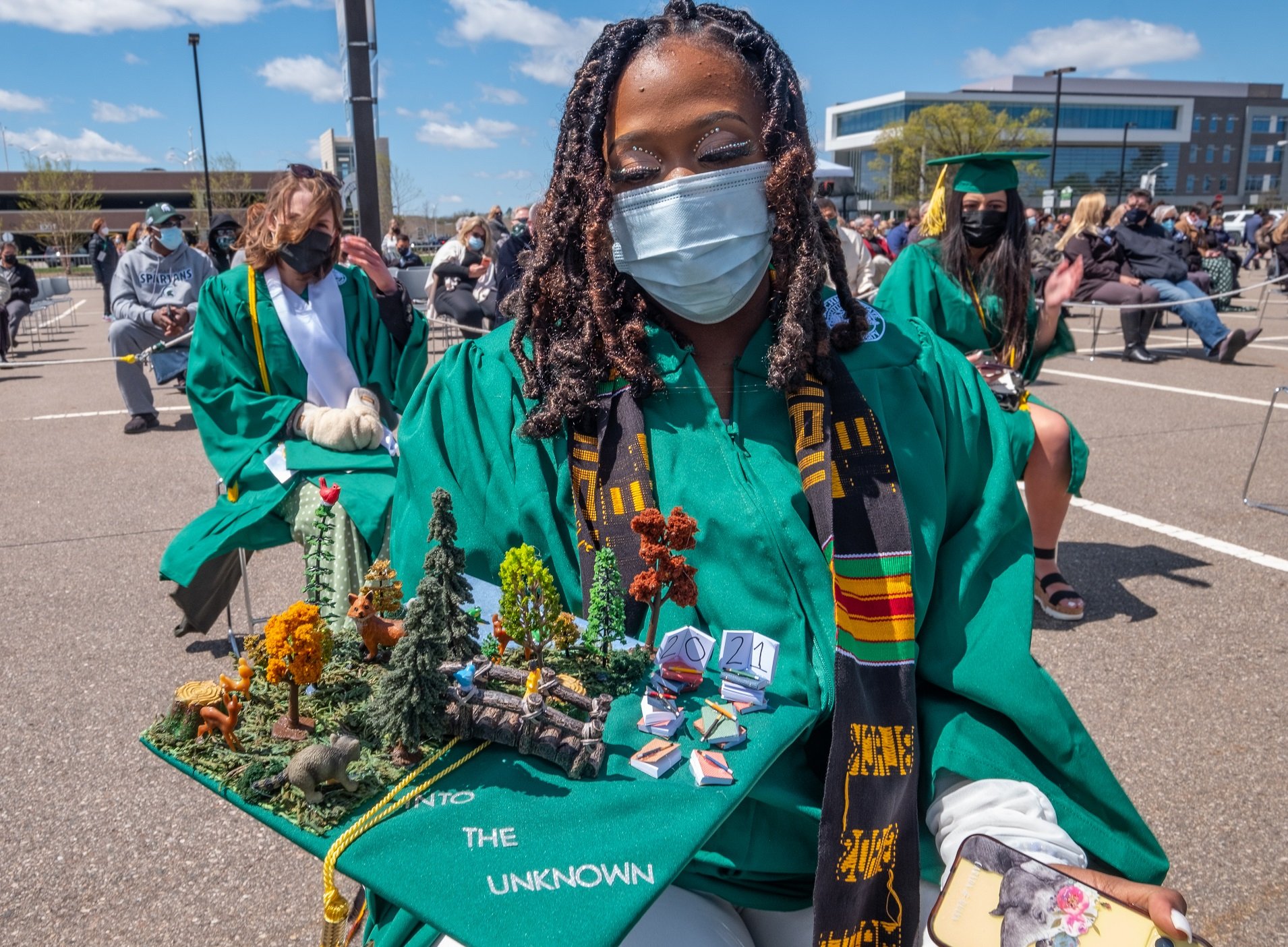 Black woman in green commencement robe sitting in a parking lot holds a decorated cap with a forest scene saying ?into the unknown.? Behind her are other graduates and blue sky with white clouds. 