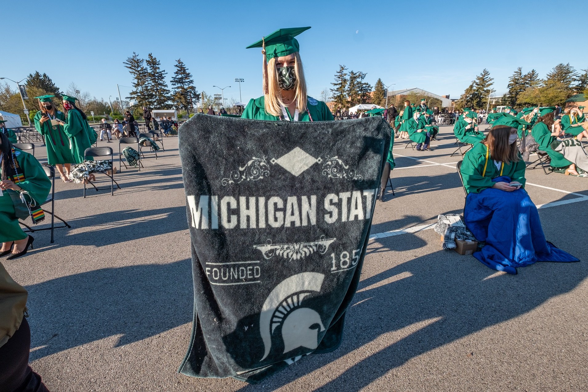 White female with blonde hair in a green cap and gown holds up a gray blanket with a white Spartan helmet and writing saying ?Michigan State Founded 1855.? She is standing in front of a chair in a parking lot with other graduates visible against blue sky. 