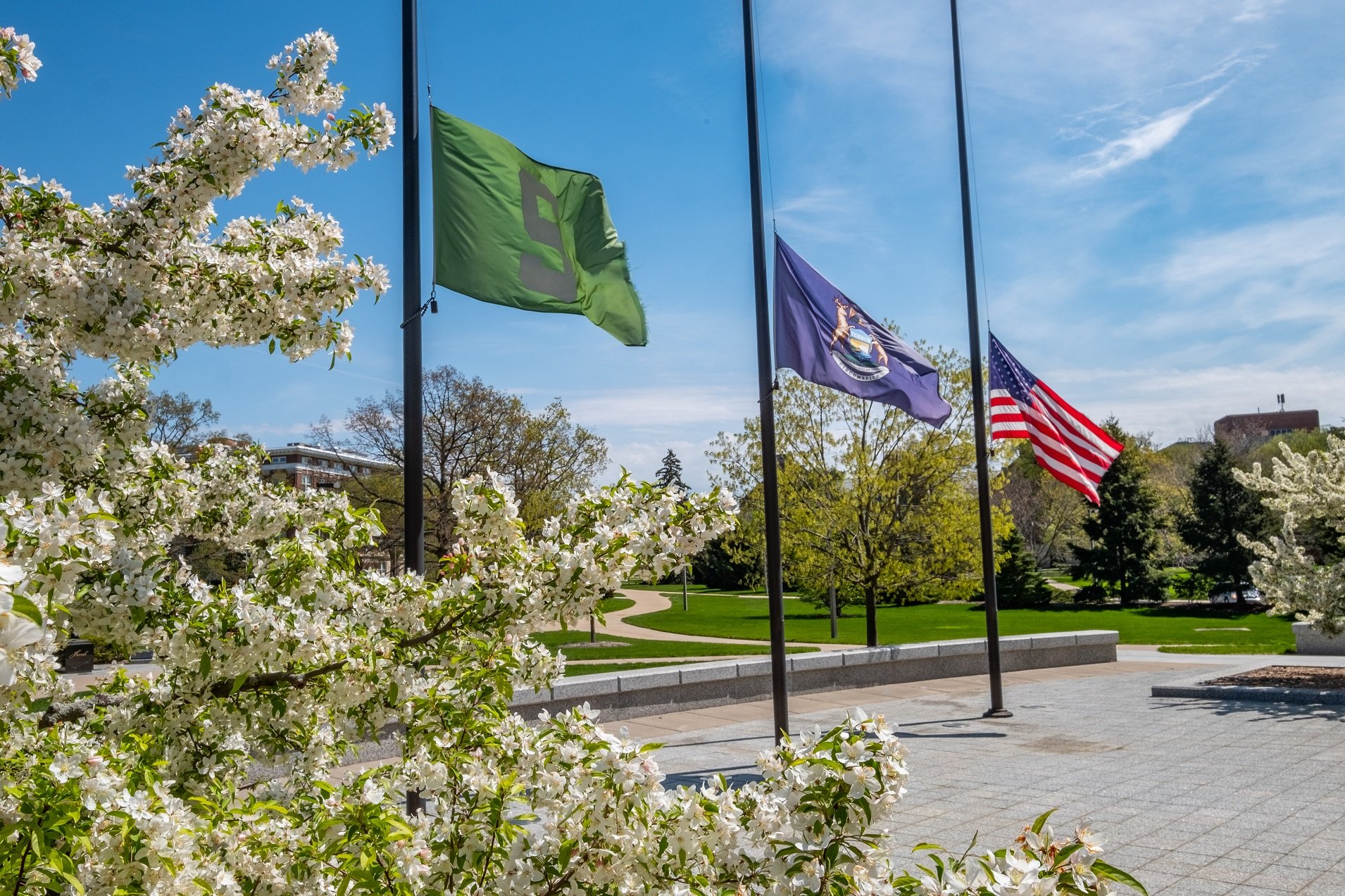 From left to right the MSU flag, State of Michigan flag and American flag hanging on flag poles unfurled in the wind against a blue sky with flowers in the foreground. All three are flying at half-mast in front of the MSU administration building. 
