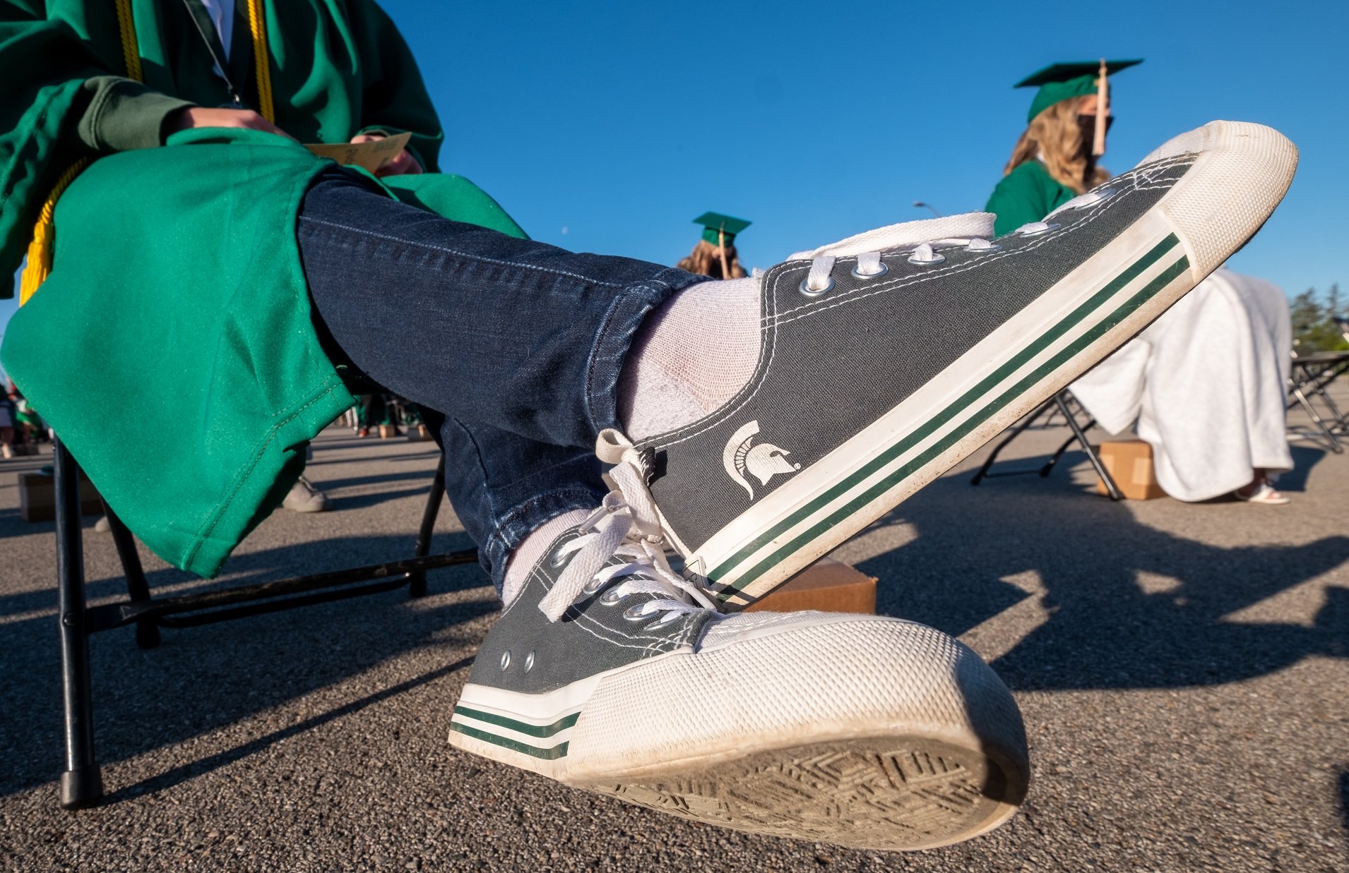 Close up view of a pair of Converse sneakers with white and green trim and white laces. The body of the sneakers is gray with a white Spartan helmet. The person wearing the shoes is sitting and also wearing jeans, white socks and a green graduation gown. 