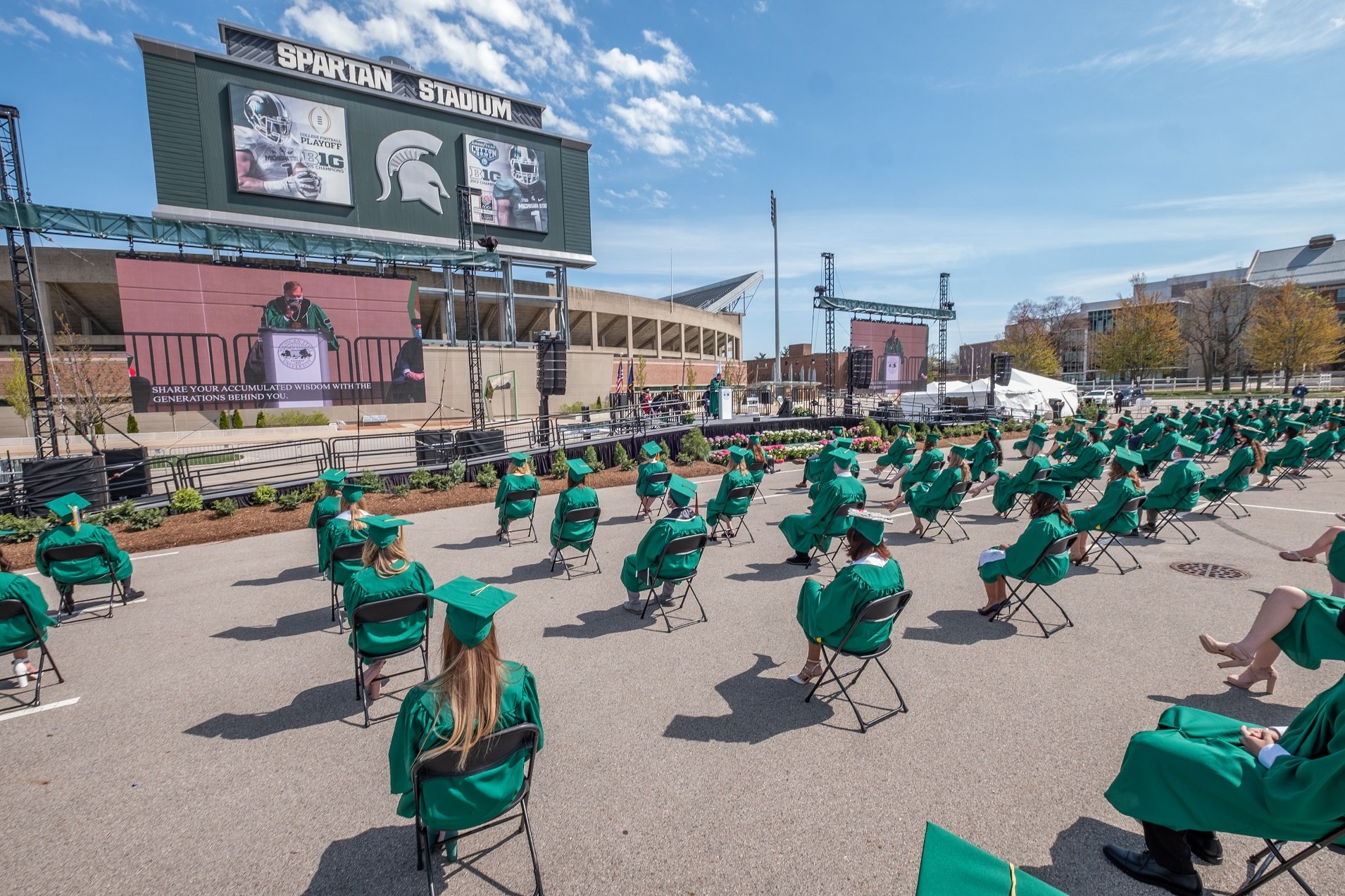 High angle view of people in green caps and gowns sitting in rows of chairs spread apart in the Spartan Stadium parking lot. The south end of the stadium is visible against a blue sky. MSU President Stanley can be seen on a large screen as he stands behind a podium in a green graduate gown. 