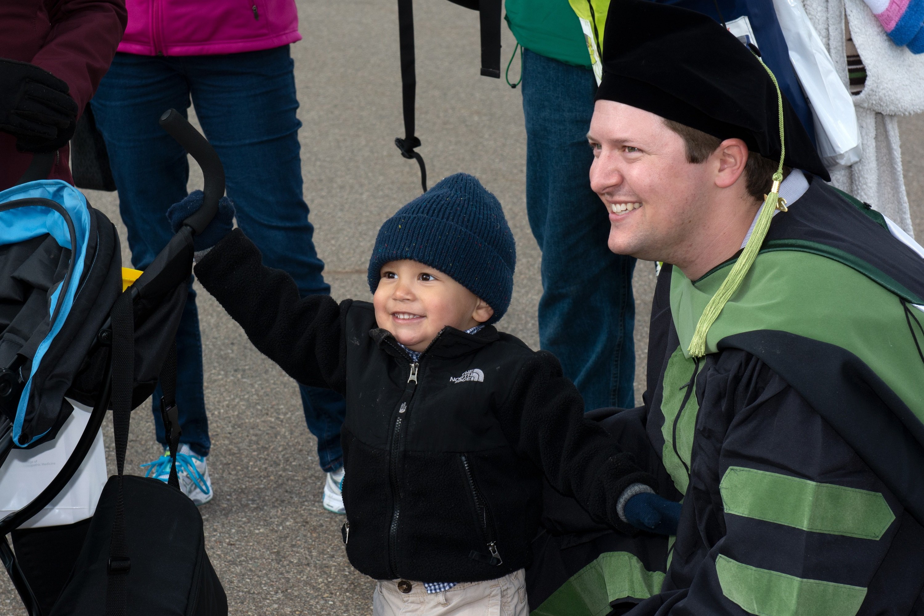 White male in a black graduation robe and hat with green trim kneels next to a toddler. Both are smiling and looking to the left.