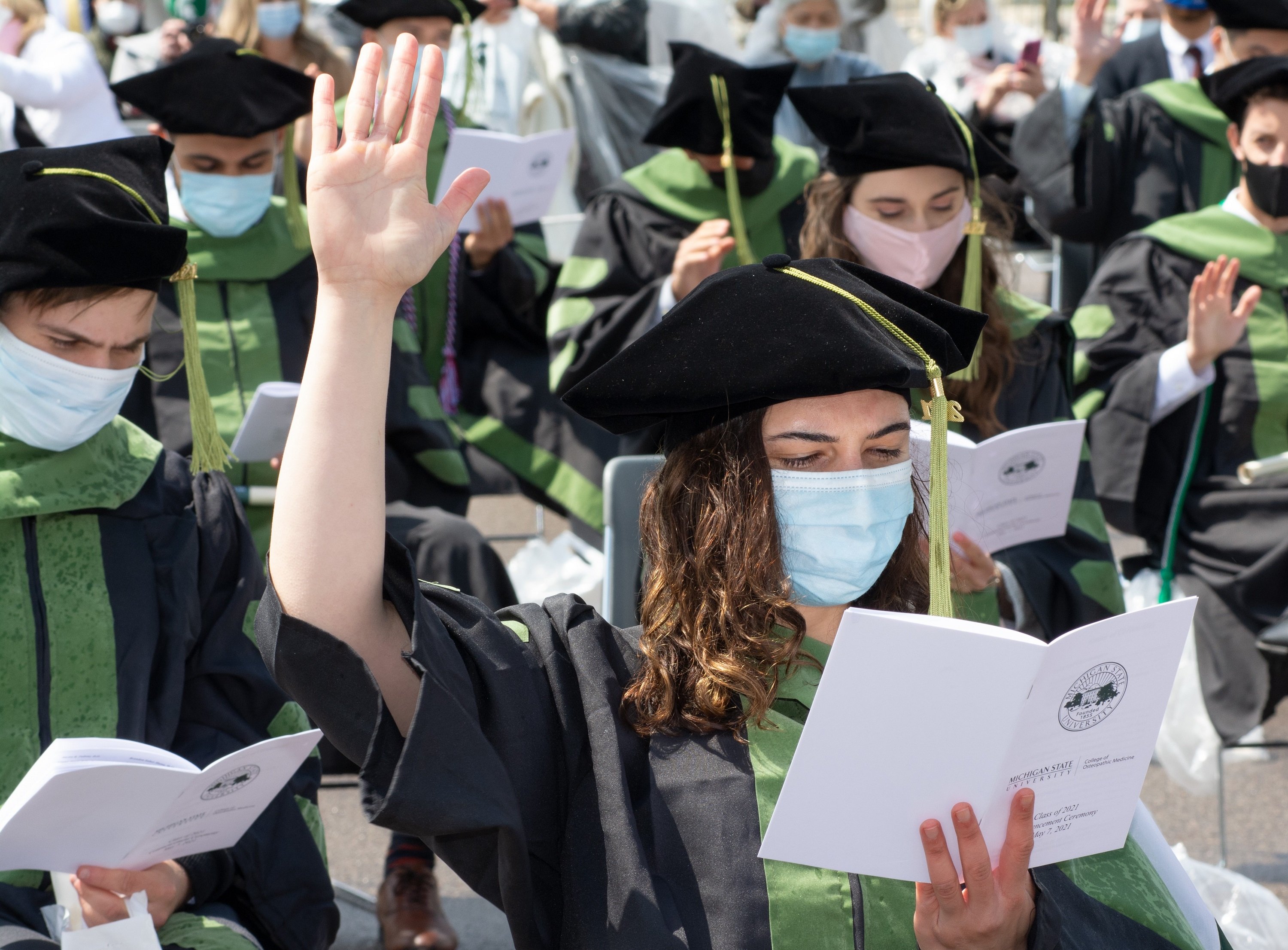 A white female in a black graduation robe and hat is prominently shown. She is raising her right hand and reading from a white bulletin. Other graduates are visible behind her.