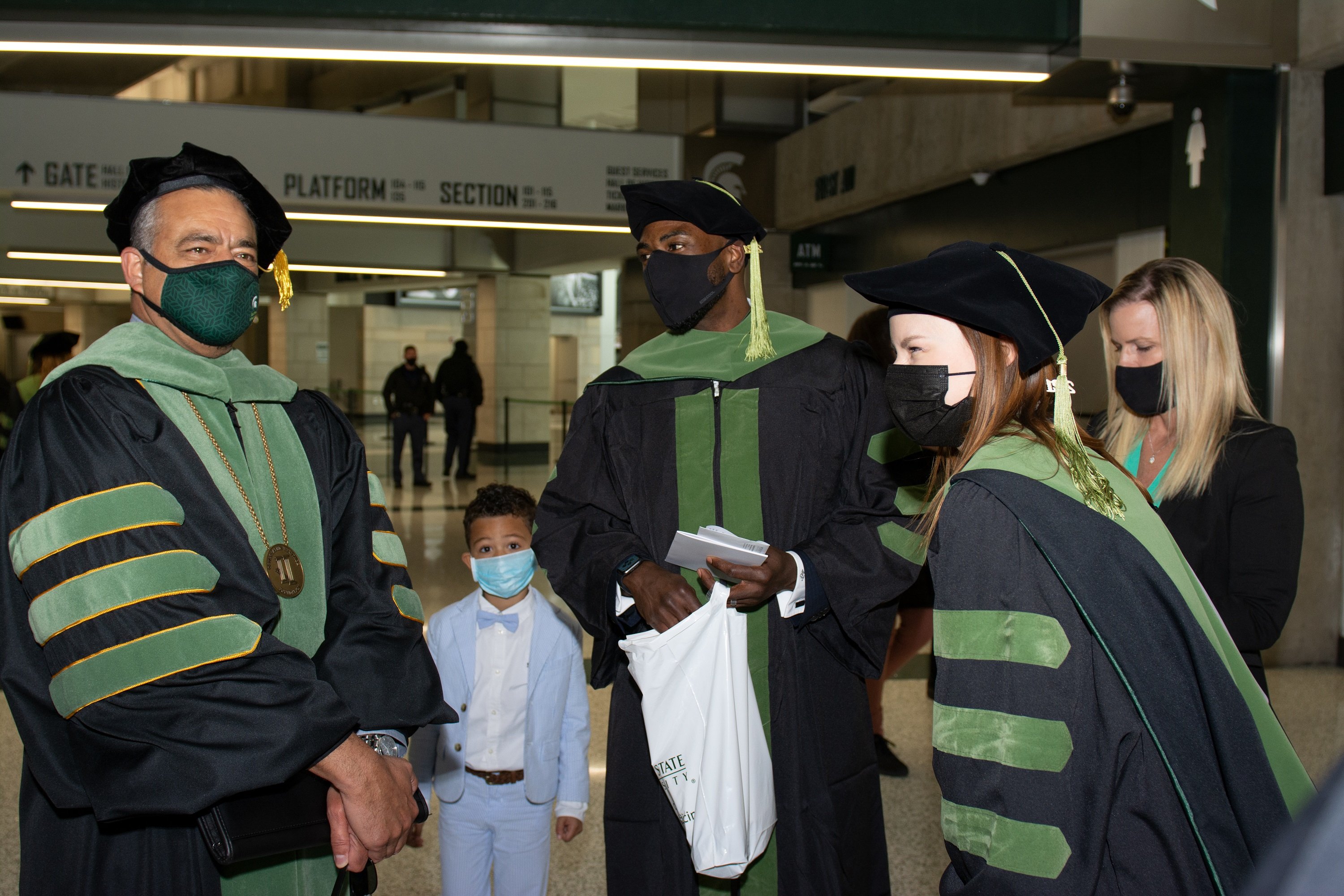 A white male, Black male, Black toddler and two white females stand on the concourse of the Breslin Center. One of the white females and both males are wearing black graduation robes and hats with green trim.