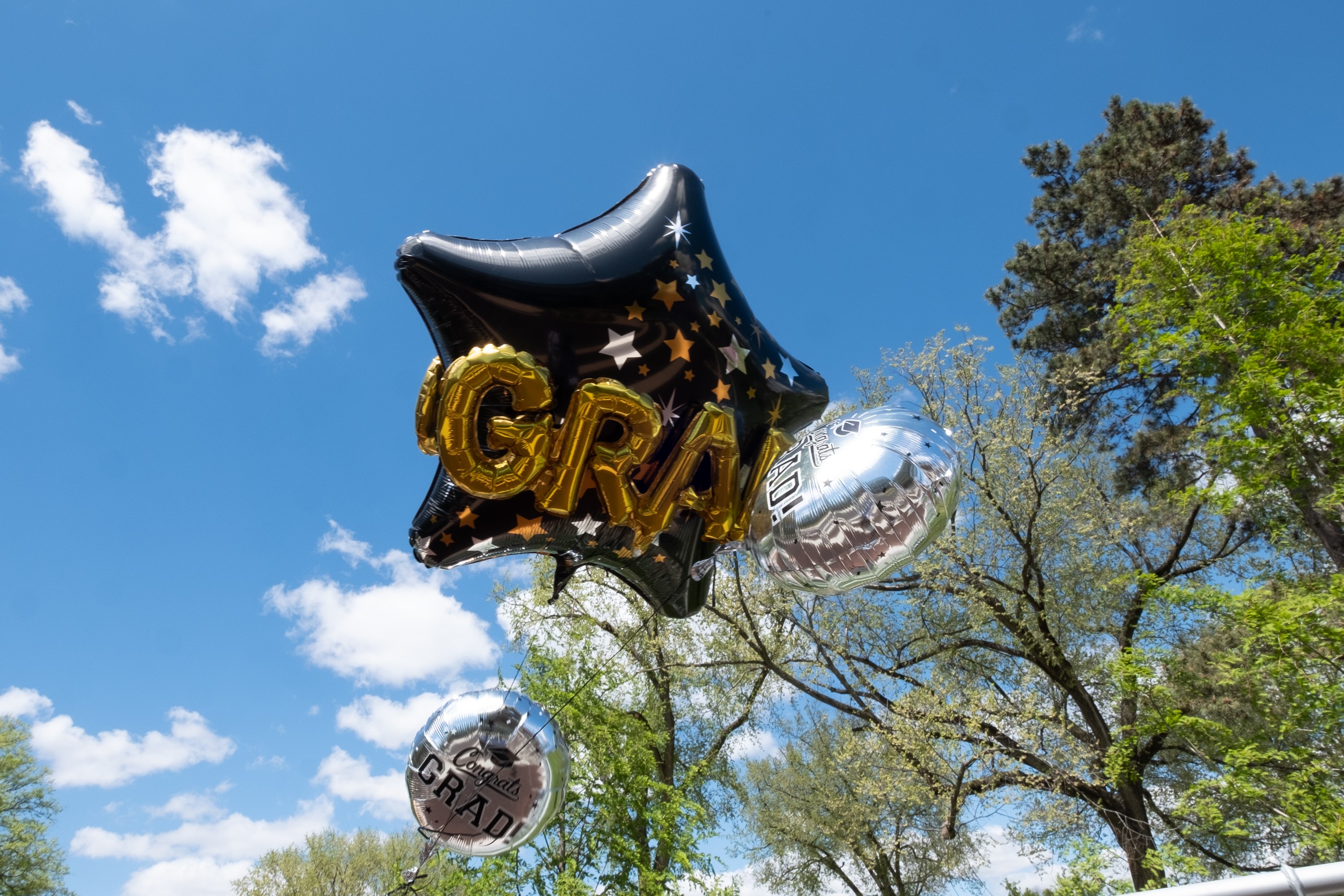 Four balloons against a blue sky with white puffy clouds and green trees. The lowest balloons on the left and right of the tie are circular, silver and shiny. The largest balloon in the center is black and shaped like a star. In front of that a gold balloon in the shape of letters spelling ?grad.?