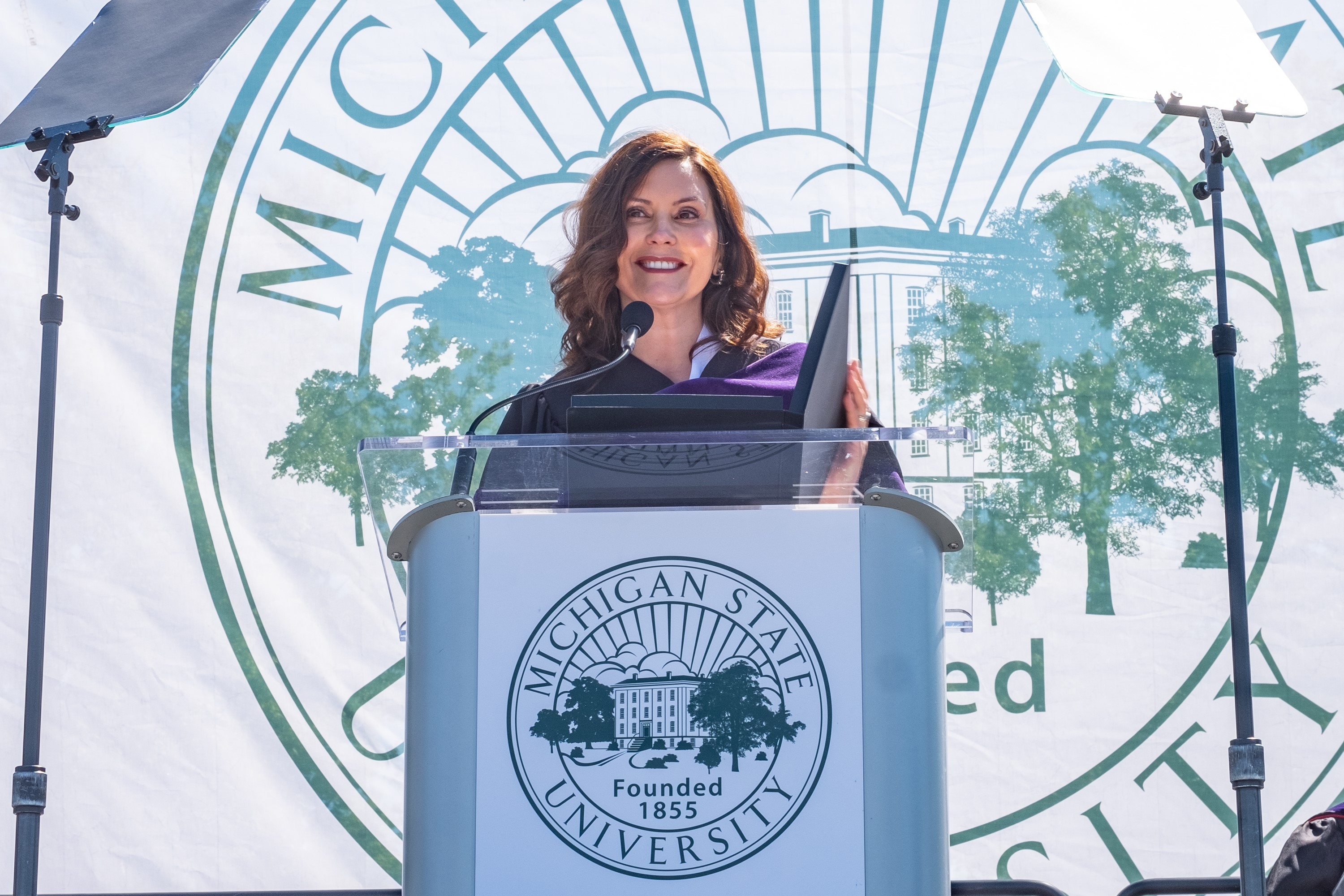 Michigan Governor Gretchen Witmer stands behind a podium with the MSU seal on the front. A larger version of the seal hangs behind her on a white sheet.