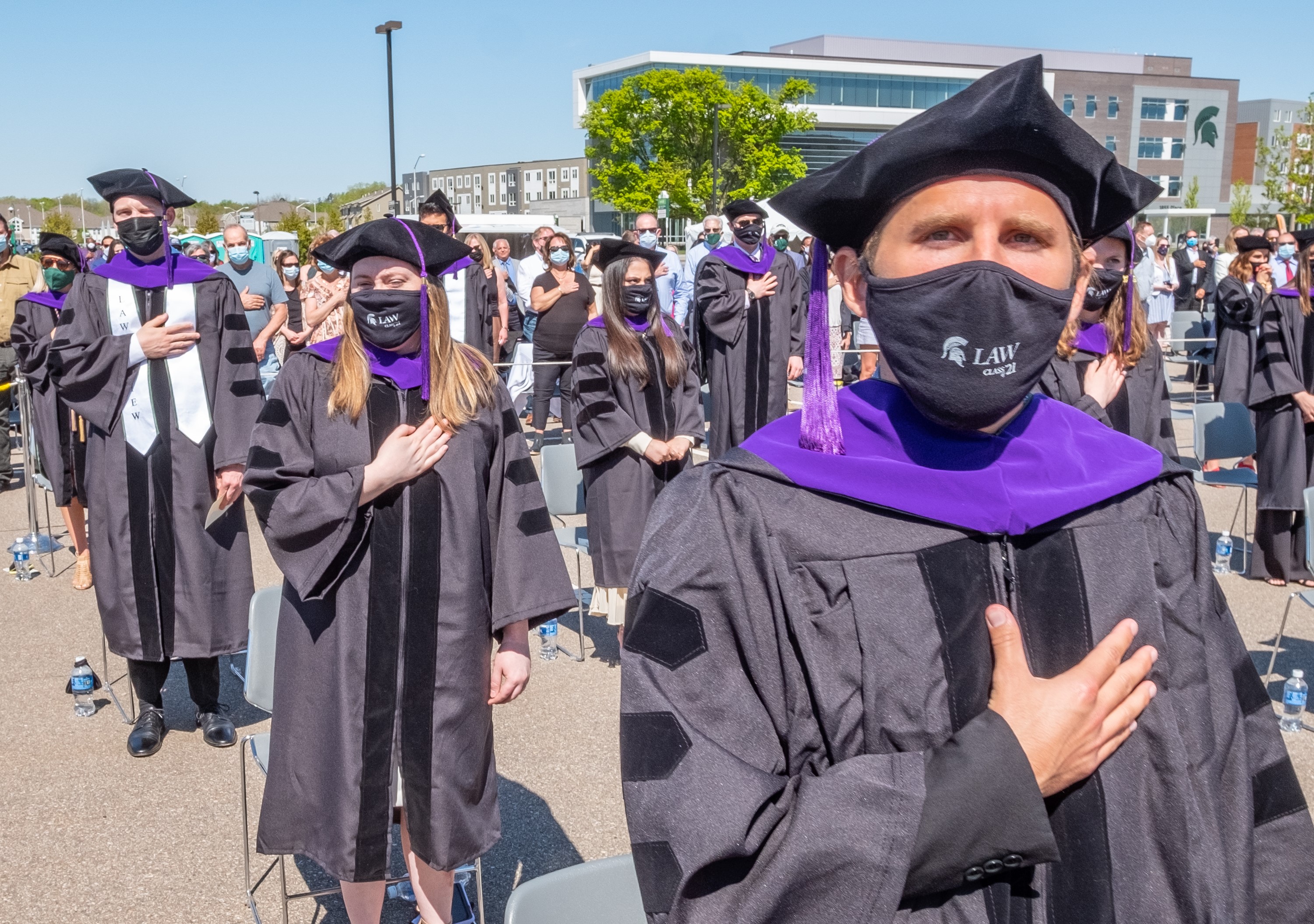 A white male in a black graduation robe and hat with a purple stole is prominently shown. He is standing in a parking lot with his right hand across his heart. Other graduates in similar clothing and positions are seen behind him.