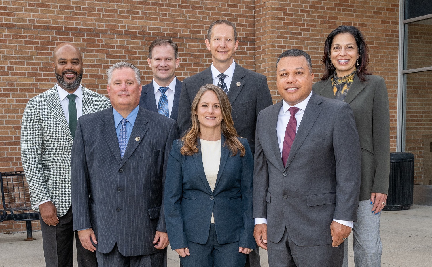 Five men and two women in suits standing in two rows in front of a red brick wall.