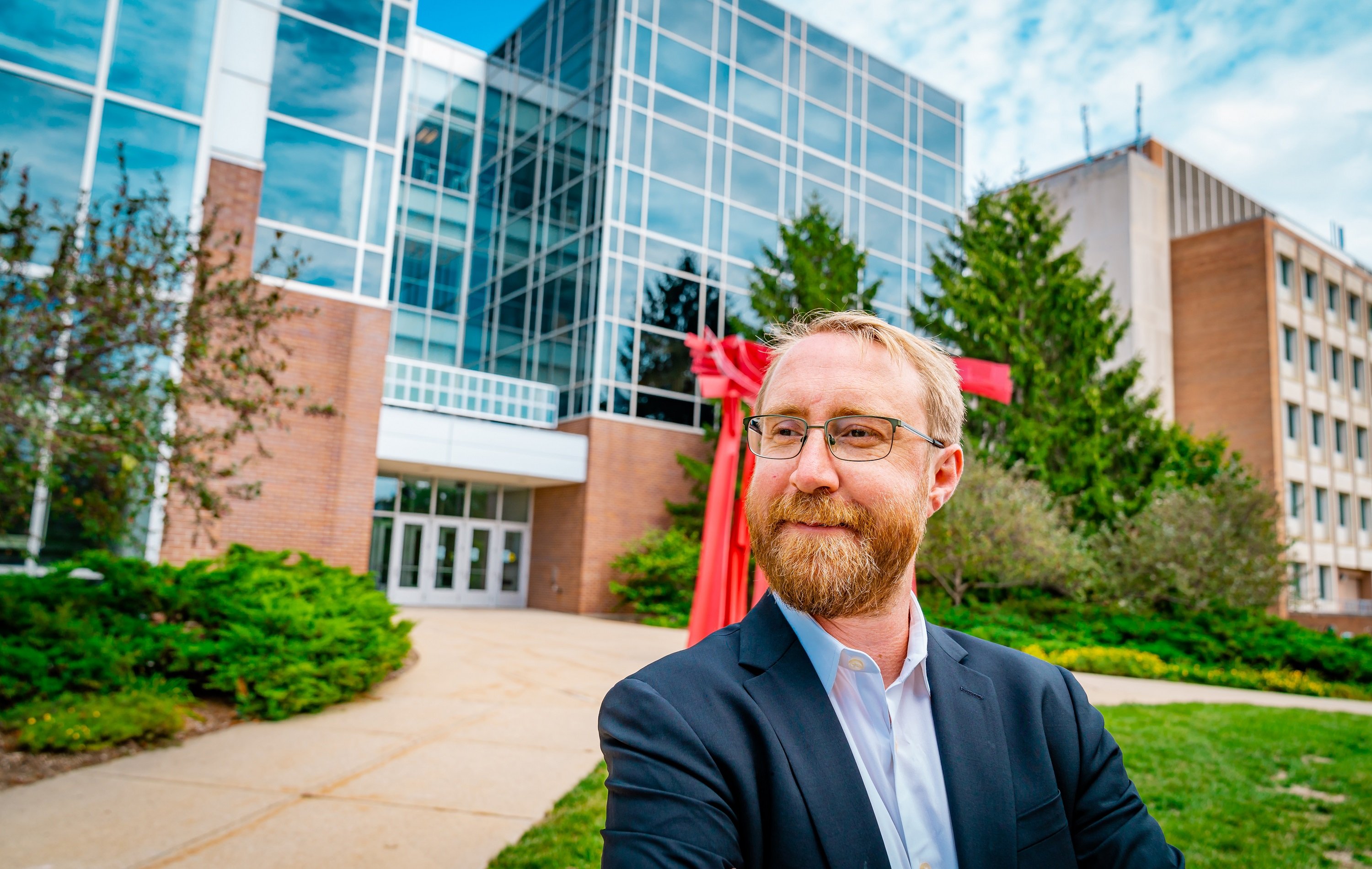 White man shown from the shoulders up in front of the Biomedical and Physical Sciences Building. He has a beard, is wearing glasses, a blue collard shirt and blue blazer. 