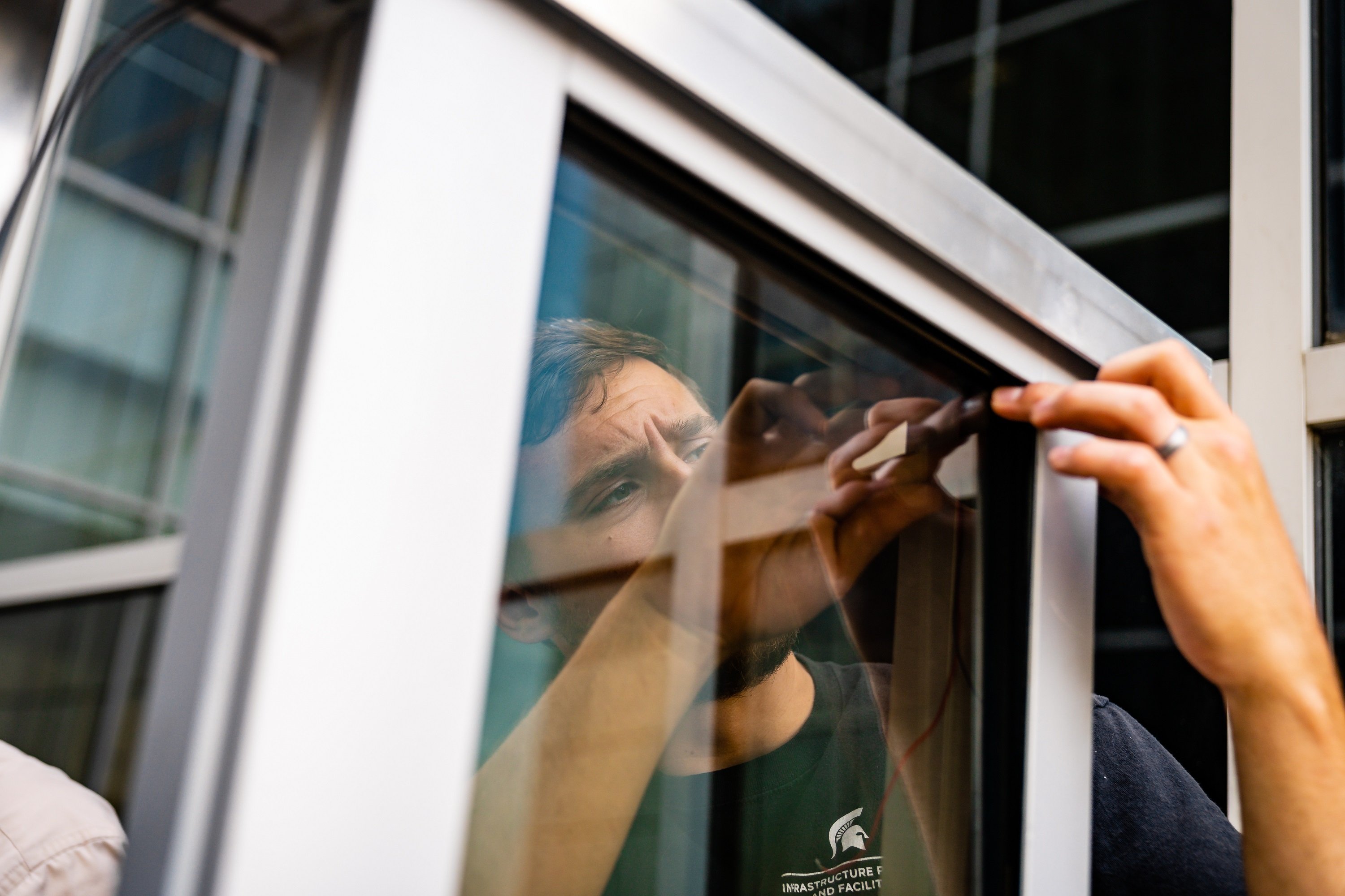 White male in a black MSU IPF t-shirt is behind a piece of glass with his hands on either side. 