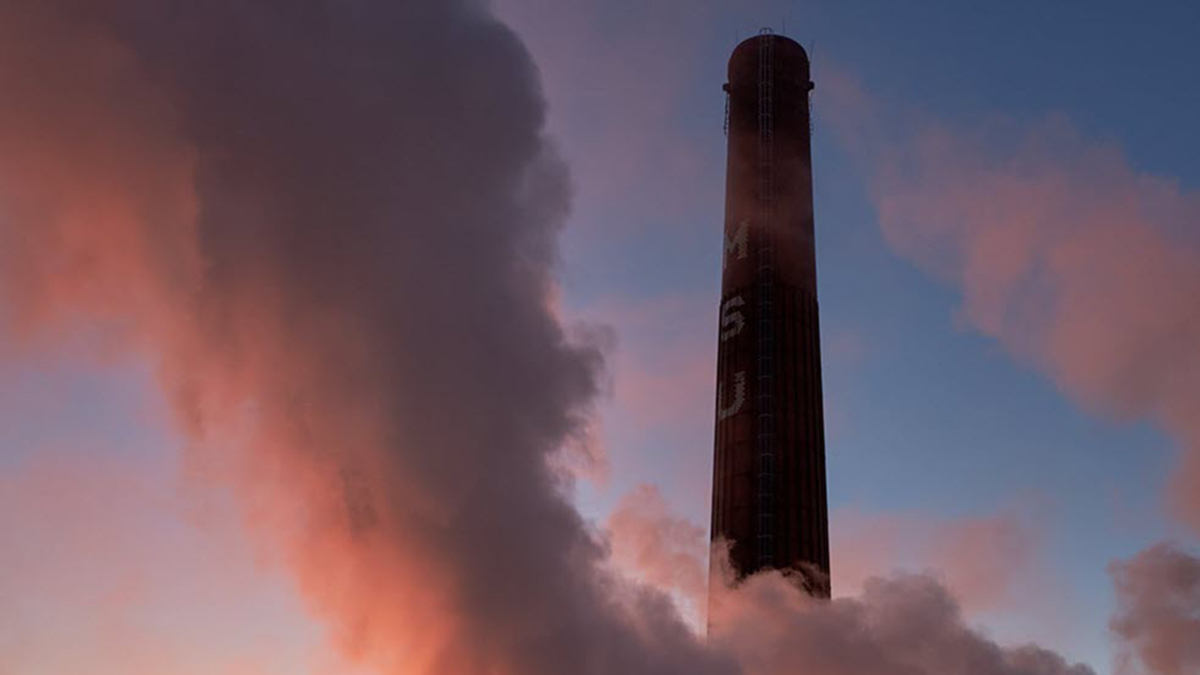 White steam rises against a blue sky with a brick smokestack rising through the photo with white letters that read