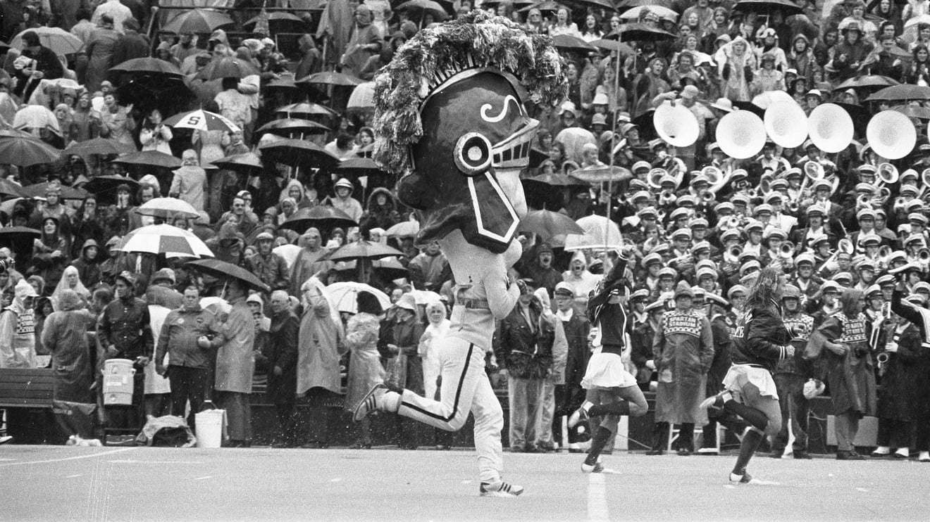Old version of Sparty running on the field with cheerleaders during a football game, 1973