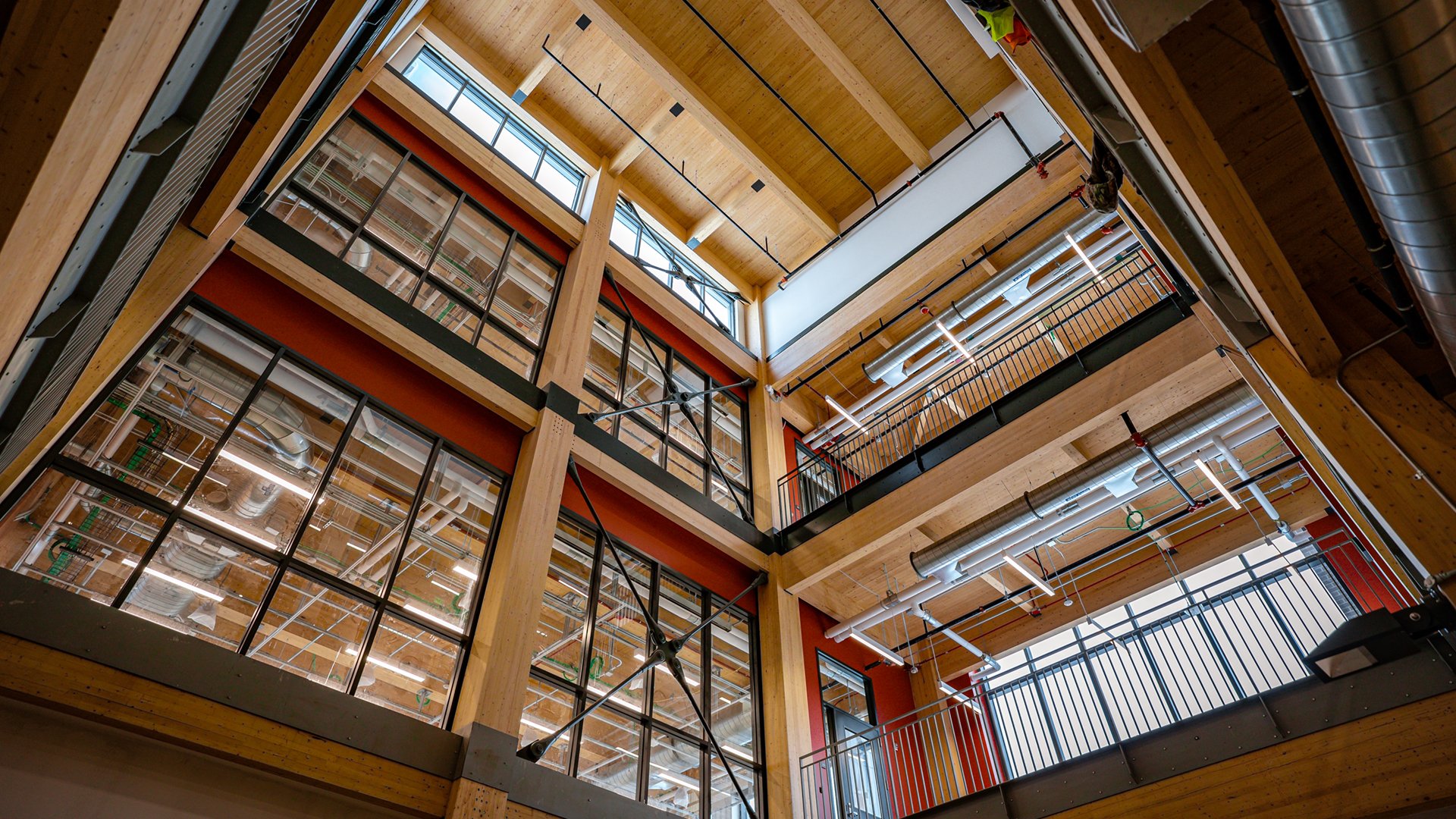 A photograph shows a view from the ground level, looking upward in a lobby of the new STEM Teaching and Learning Facility. The view shows how glass, steel and mass timber come together to give the building an open, airy esthetic that is part cozy lodge and part industrial loft.