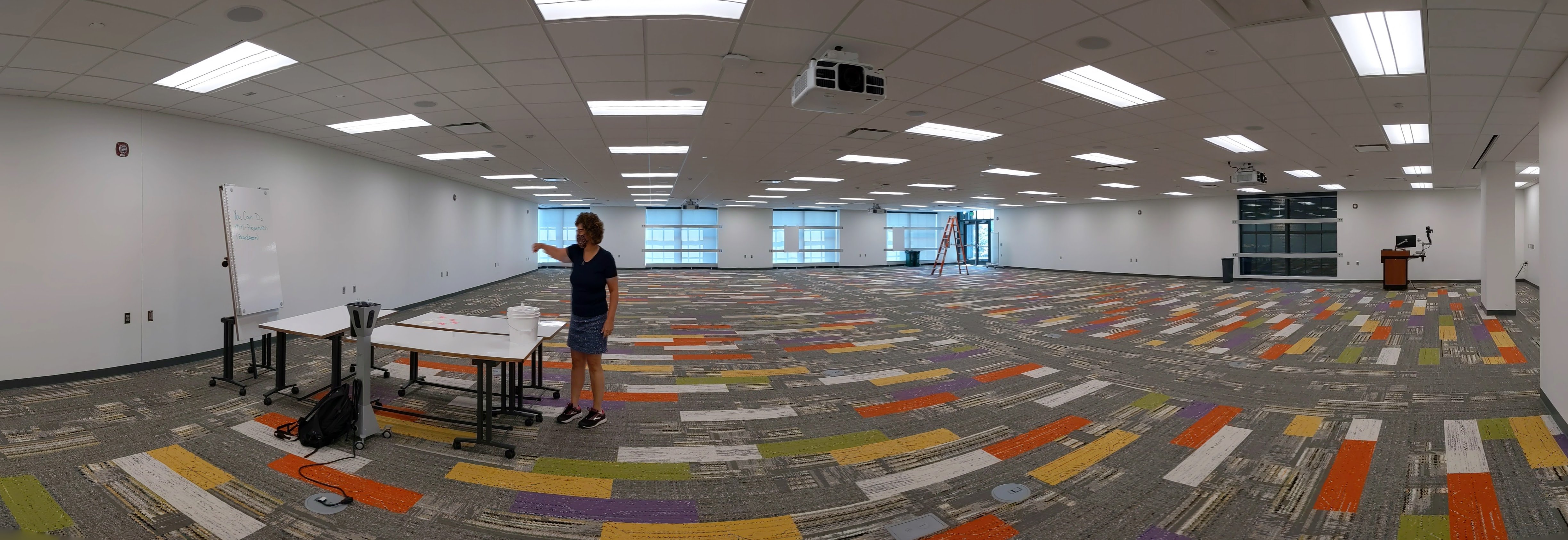 A panoramic photograph shows a large lecture hall designed for 100-plus students. Barbara Kranz, director of Facilities Planning and Space Management, stands near three tables with white boards tabletops. 