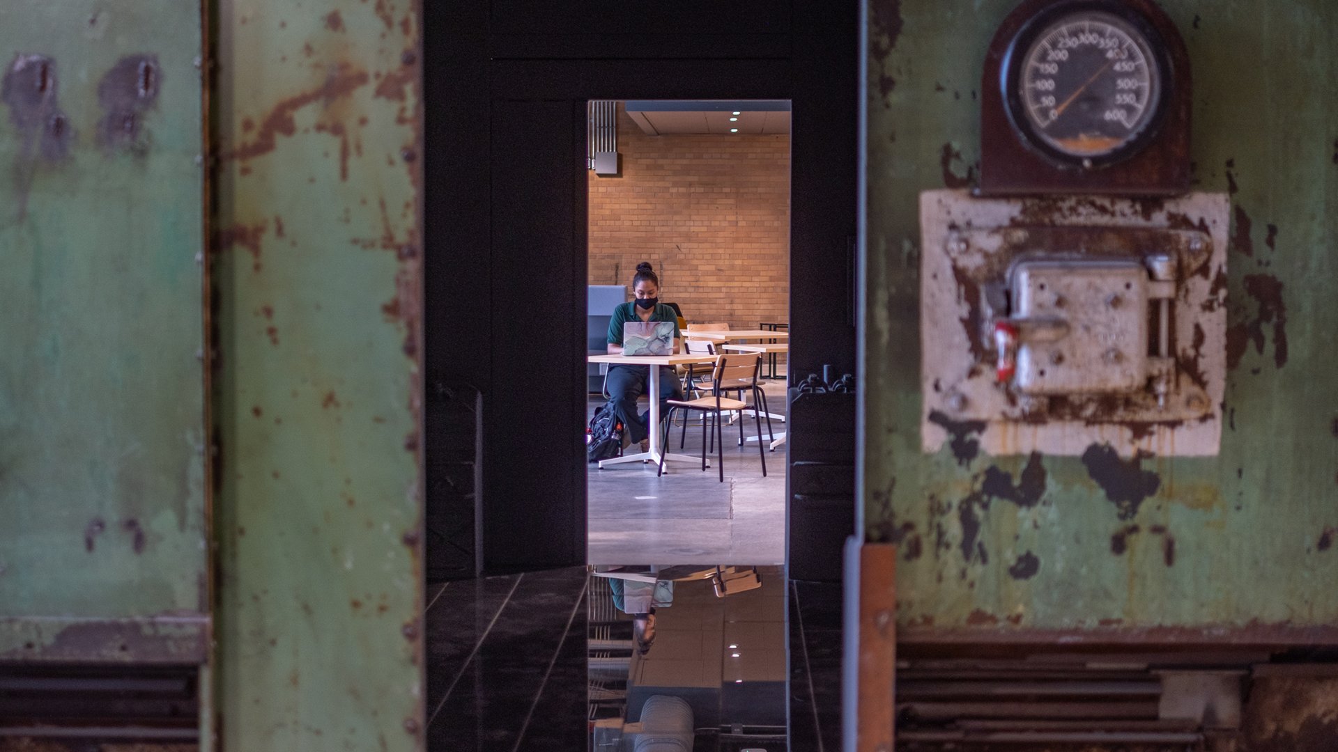 A photograph shows a student working in a study and meeting space in the new STEM Teaching and Learning Facility. The picture is taken through the original boiler from the Shaw Lane Power Plant, which has been transformed into a visual art installation.