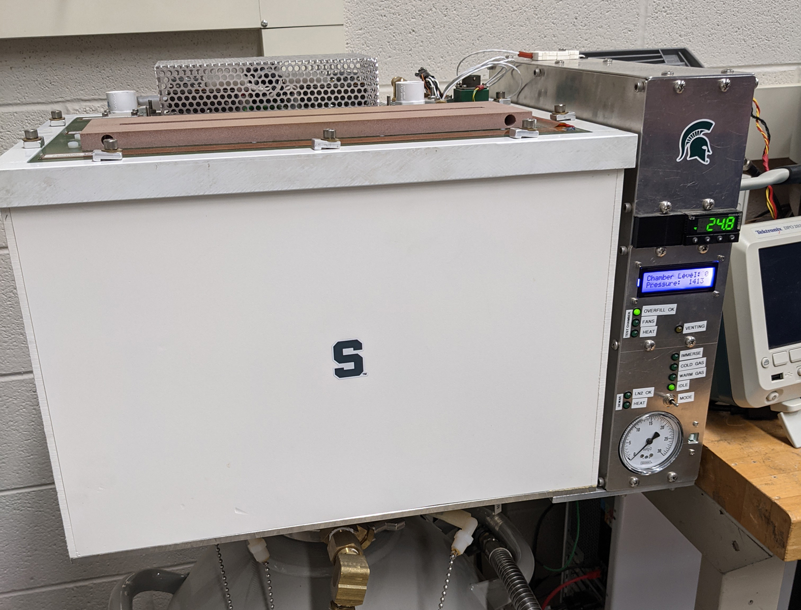 A white box with a green block “S” sticker is connected by wires to a slimmer silver box with a digital display reading “248”, an analog gauge and a green Spartan helmet.