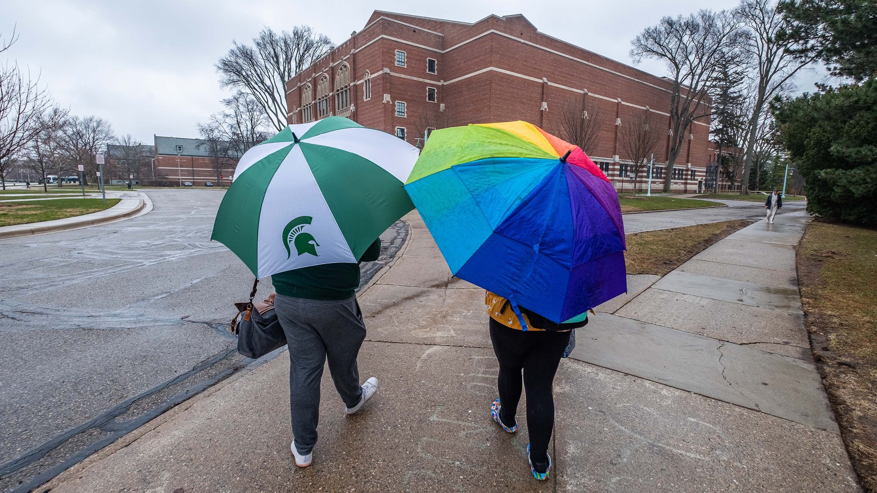 Two people walking on campus, one holding Spartan-themed umbrella, one holding rainbow umbrella