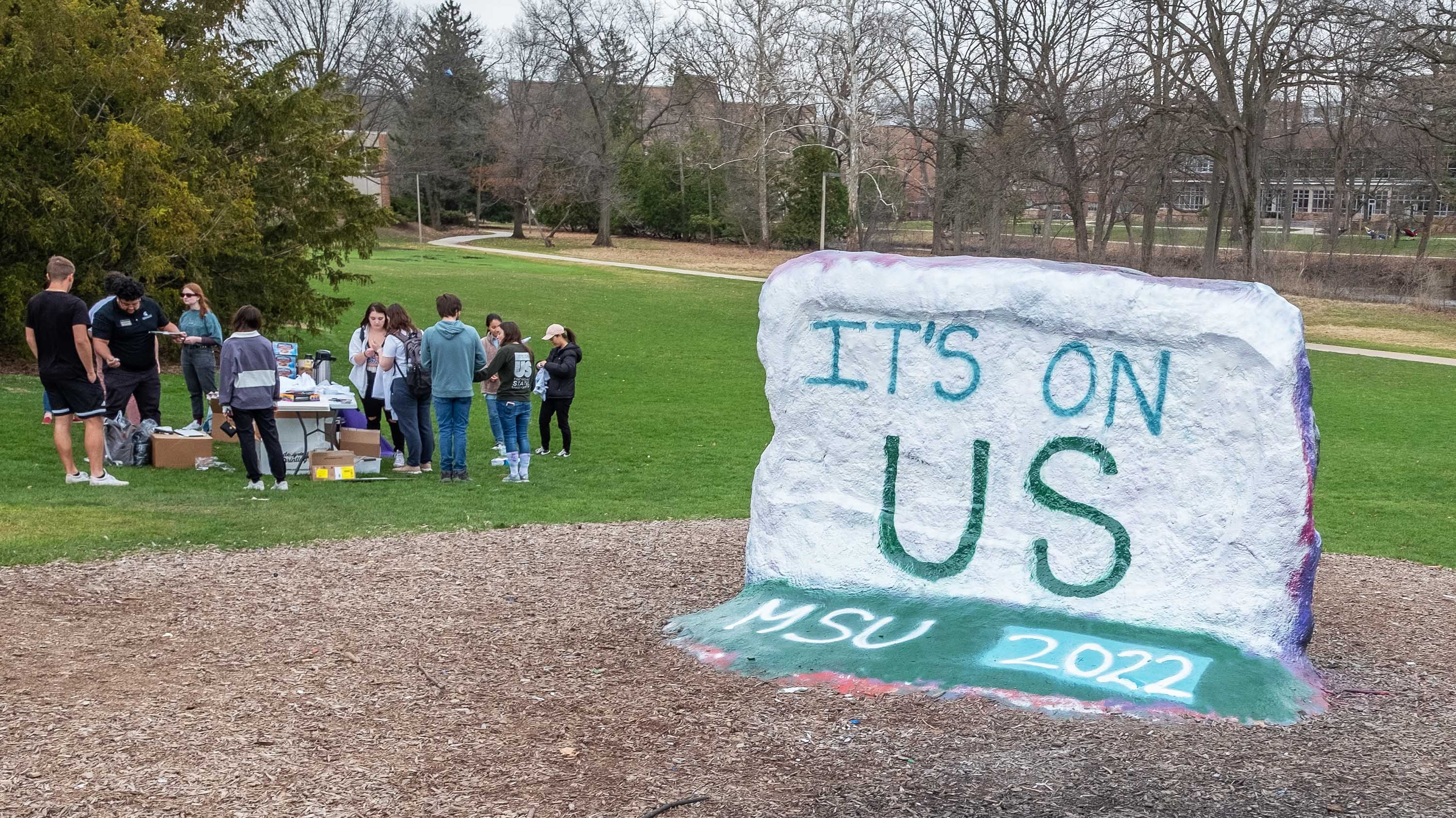 MSU Rock painted with 'It's On Us' logo, in front of students participating in tie-dye event