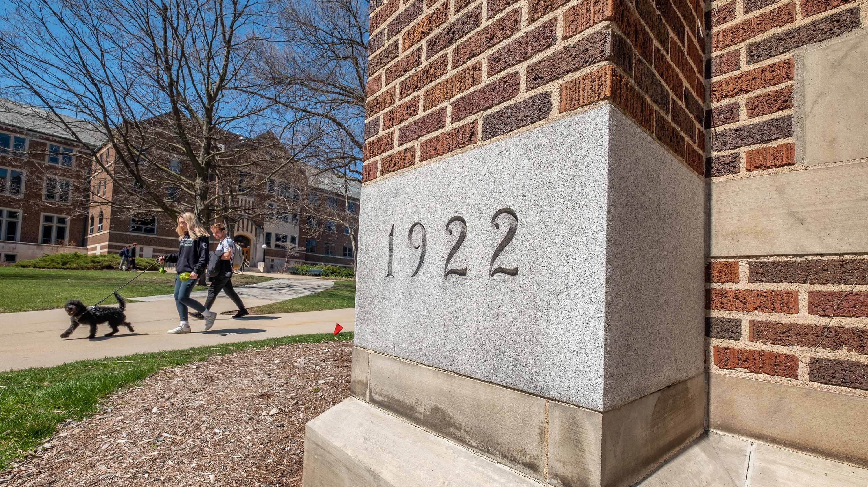 Students and dog walking past the Human Ecology corner stone, which reads '1922'