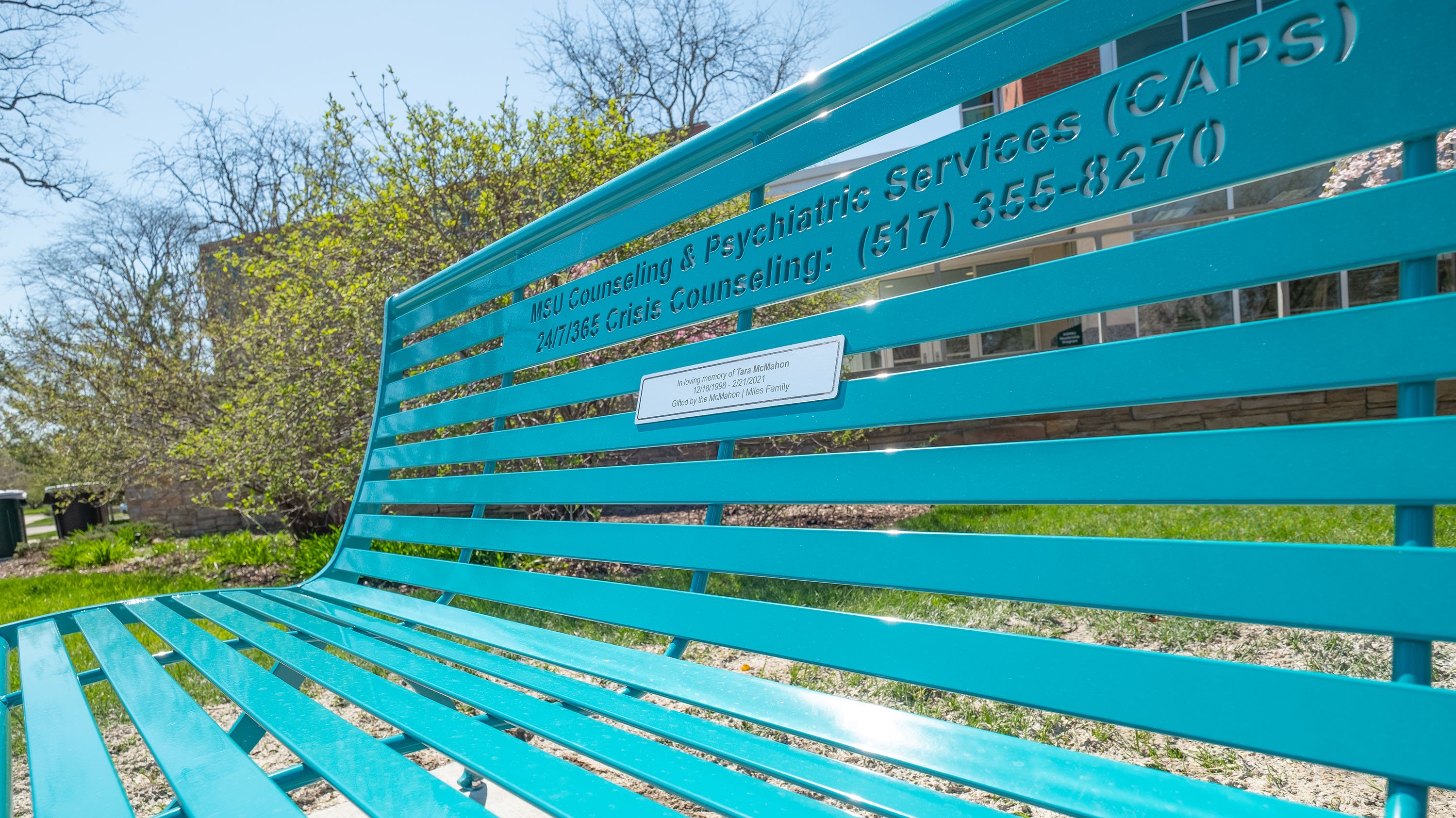 Teal bench with CAPS and crisis counseling phone numbers engraved into the back, as well as a plaque remembering Tara McMahon.