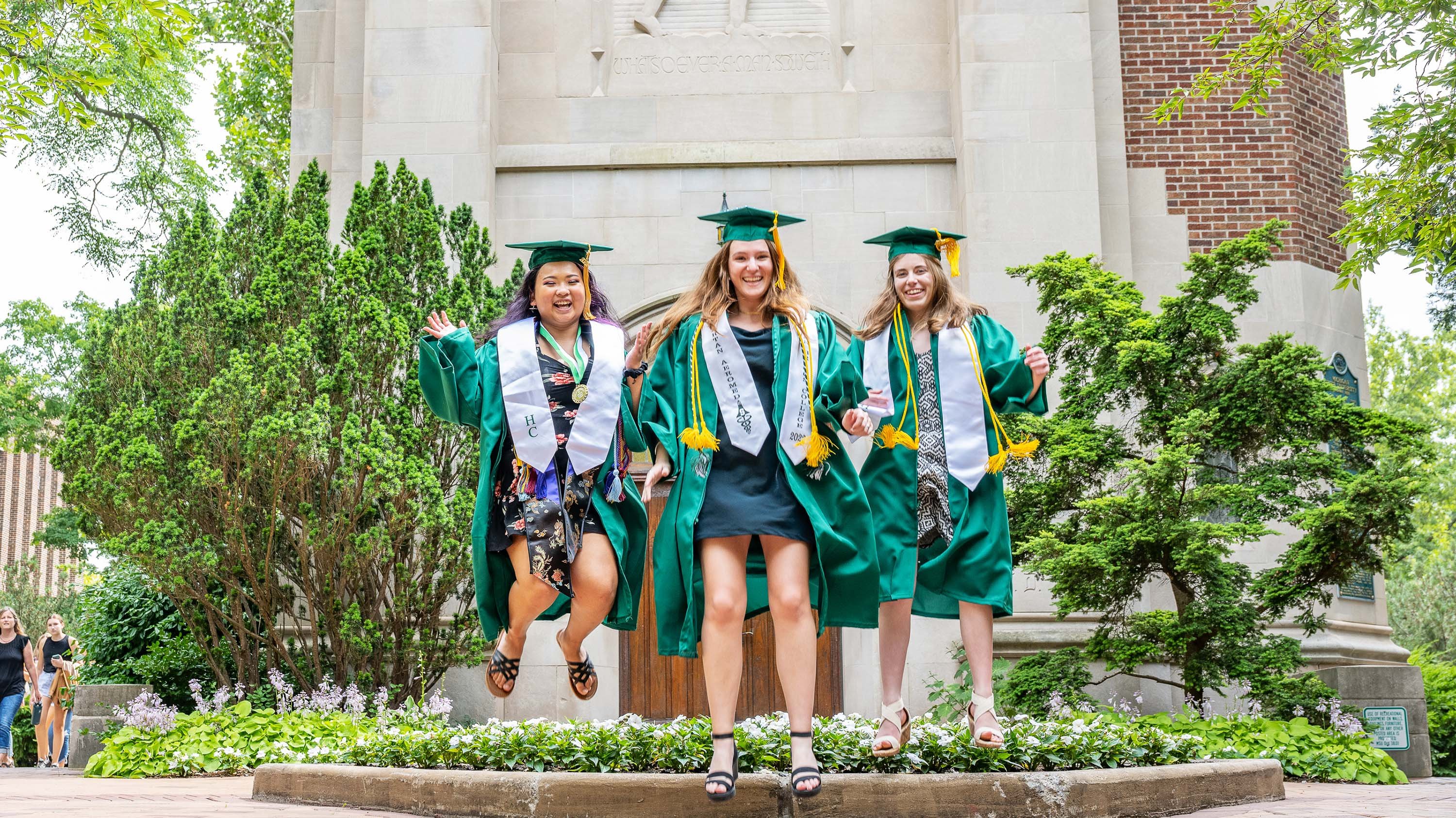 Three MSU grads in cap and gowns jumping for a photo in front of Beamont Tower