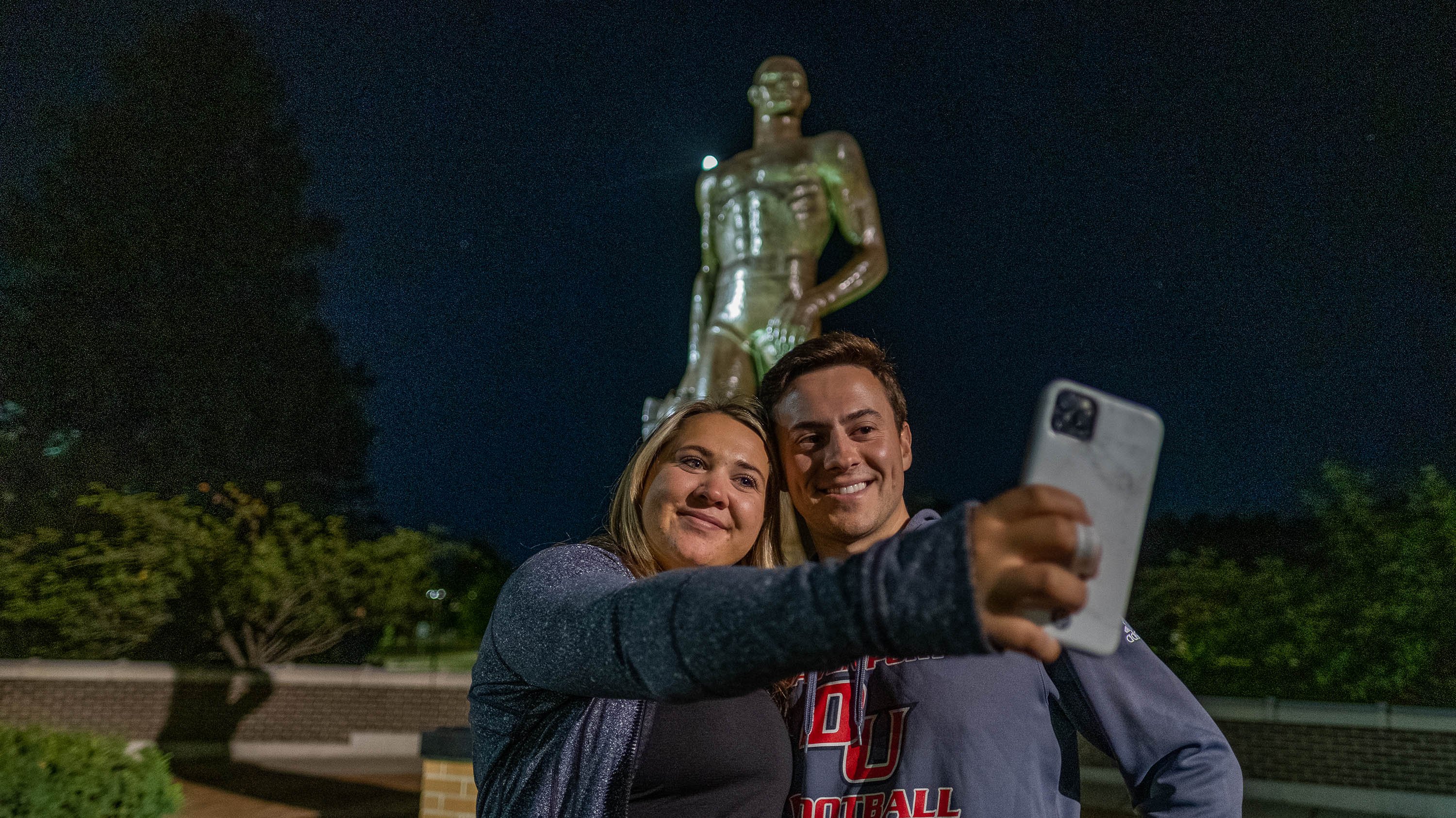 Two people taking a selfie in front of the Spartan Statue at night