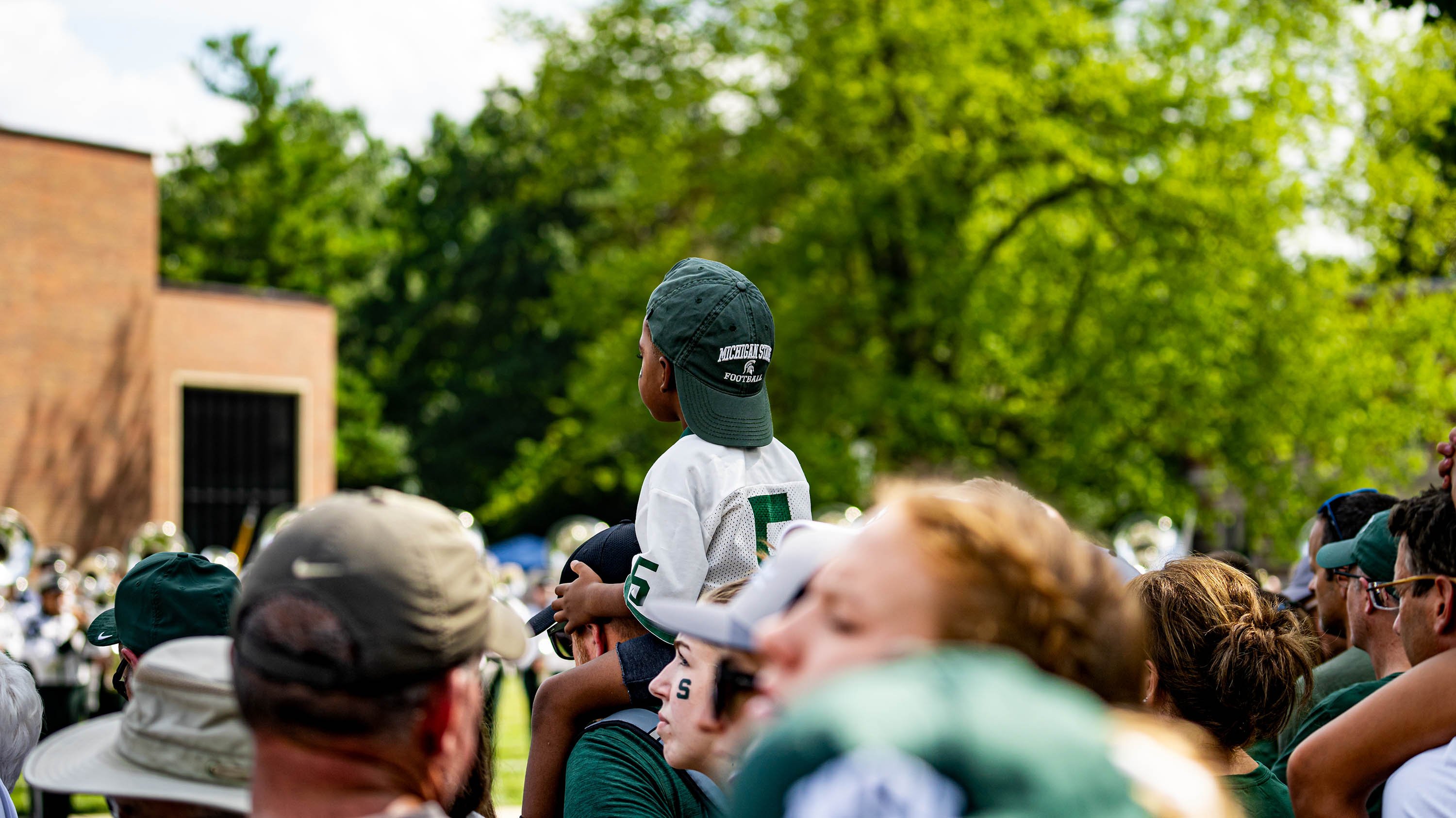 Child sitting on someone's shoulders above crowd, wearing MSU baseball cap