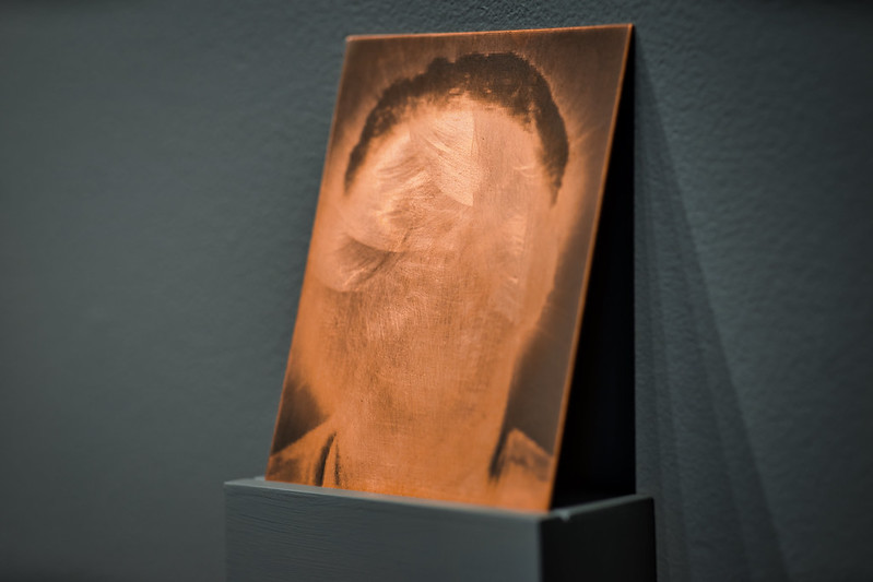 A copper-plate photogravue of a man’s mug shot rests on a wall in the “hammer without a nail exhibit.” The face, obscured by a “flash” of a faux camera, is that of Dan Paz’s father. The mug shot was purchased by Paz from a third-party criminal search website.