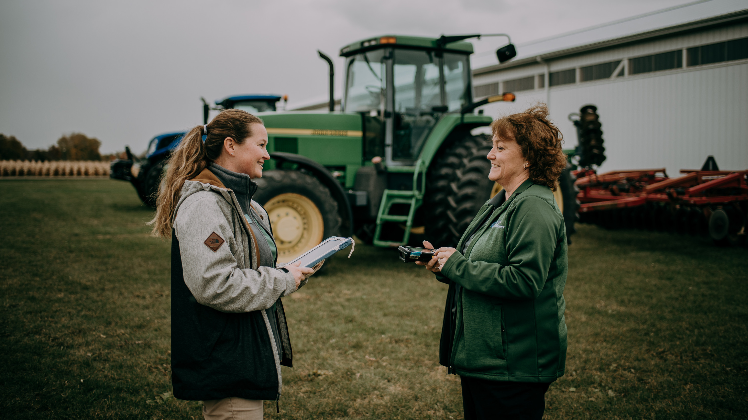 Anna Foster and Debbie Smith stand in front of a large green tractor at the Plant, Soil and Microbial Science Agronomy Farm.