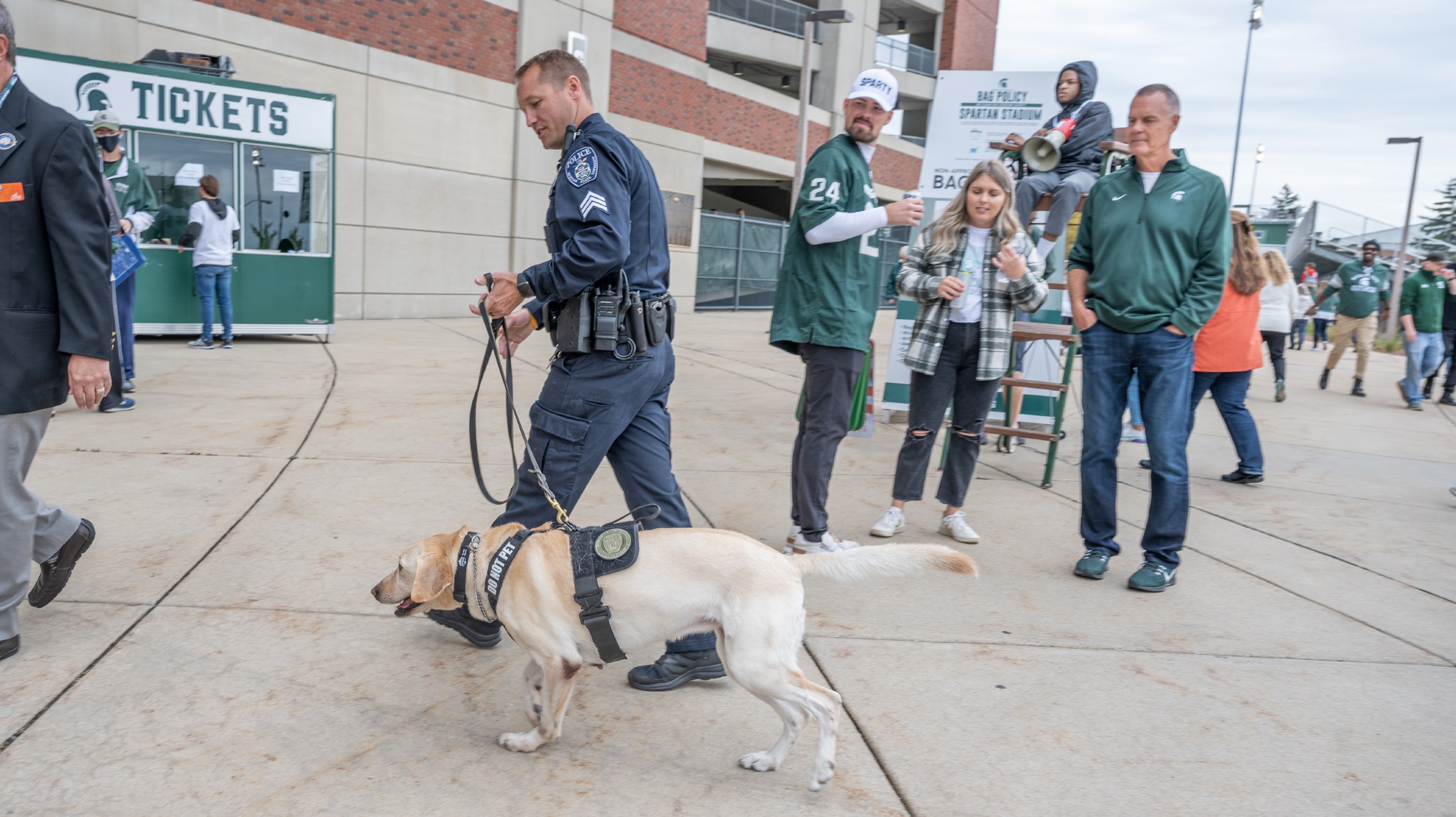 Sgt. Adam Atkinson walks with K9 Cora outside of Spartan Stadium with fans nearby.