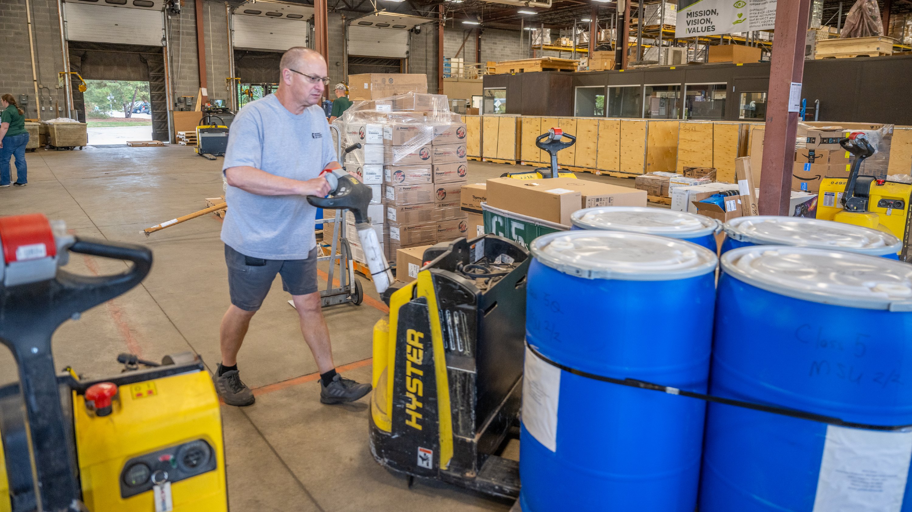 A man wearing glasses, a gray T-shirt and shorts pushes a motorized pallet jack truck loaded with blue barrels.