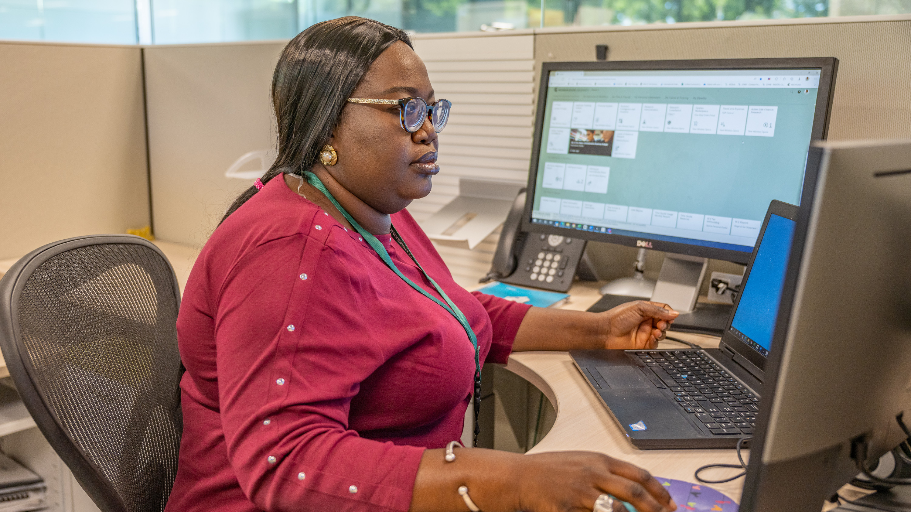 A Black woman with brown hair, wearing a pink top, sits at a desk looking at computer screens.