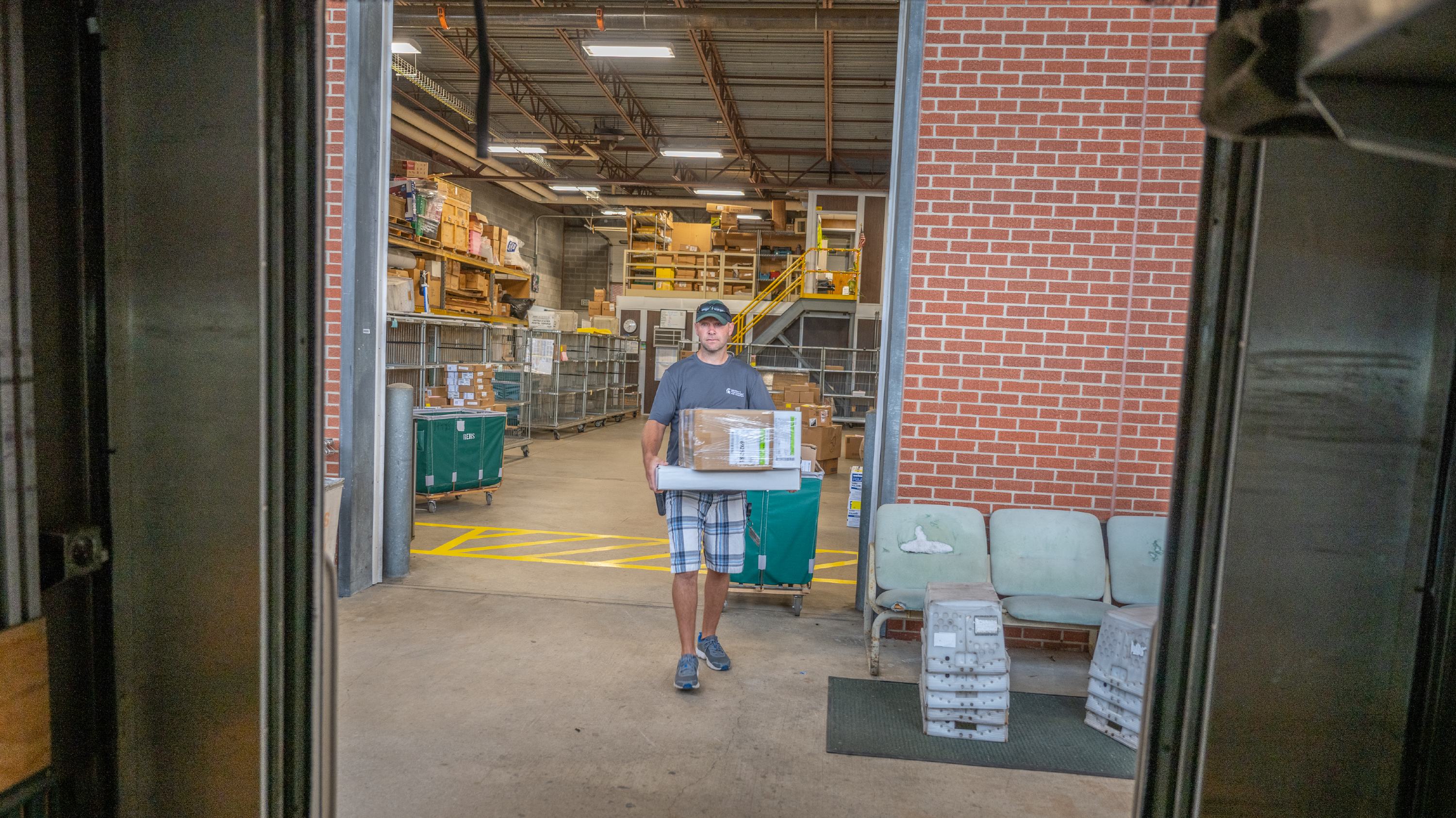 Viewed from inside the back of a truck, a man walks forward carrying three boxes. 