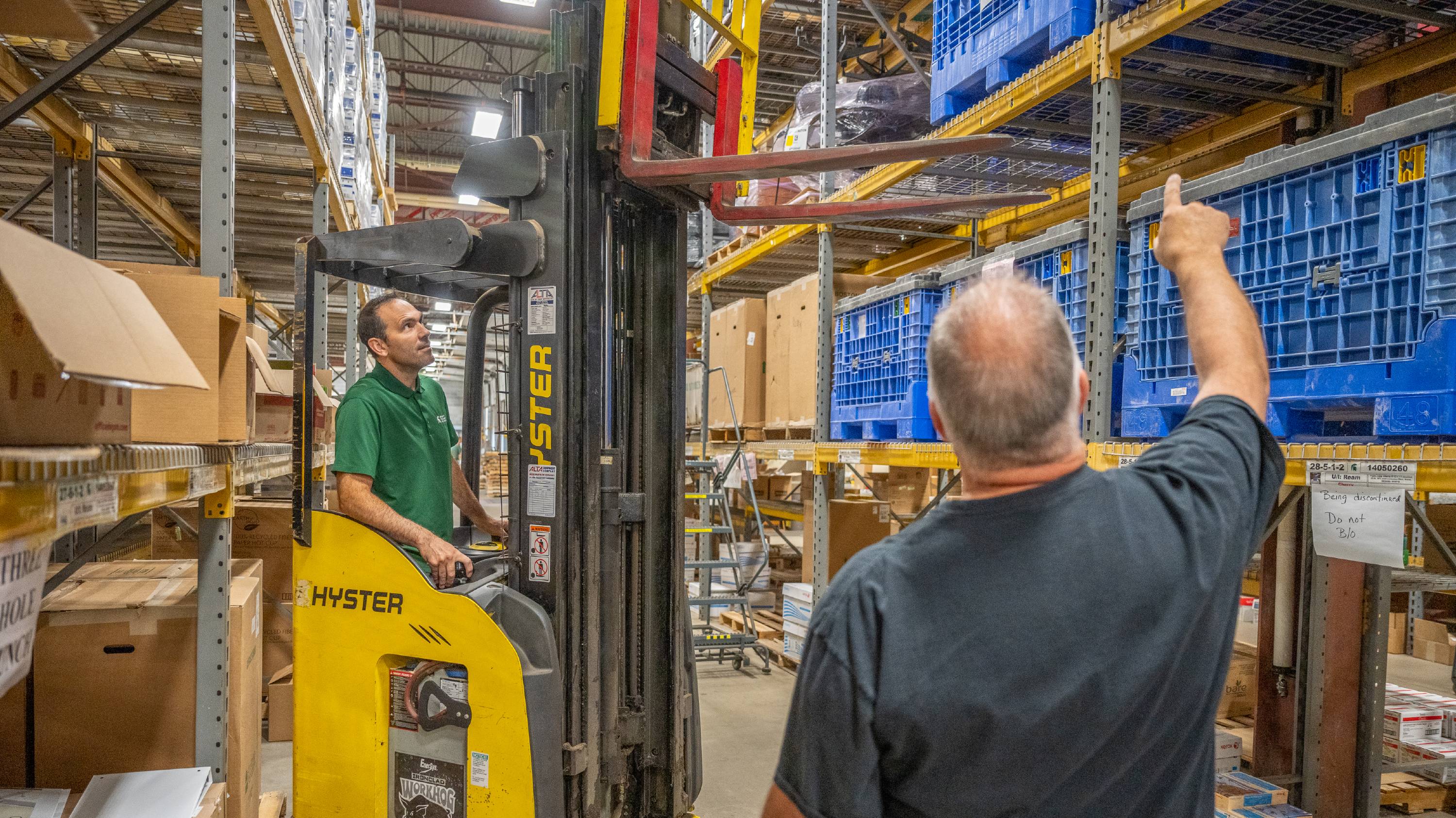 A man in a green shirt operates a forklift. A male in a gray shirt is seen from the back pointing up at storage shelves.
