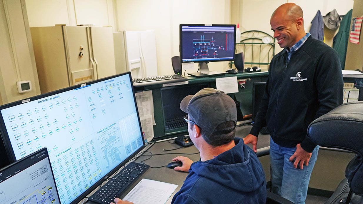 Two workers, one seated and one standing, look at computer screens.