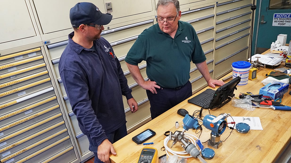 Two workers stand in front of a table with equipment and a laptop.