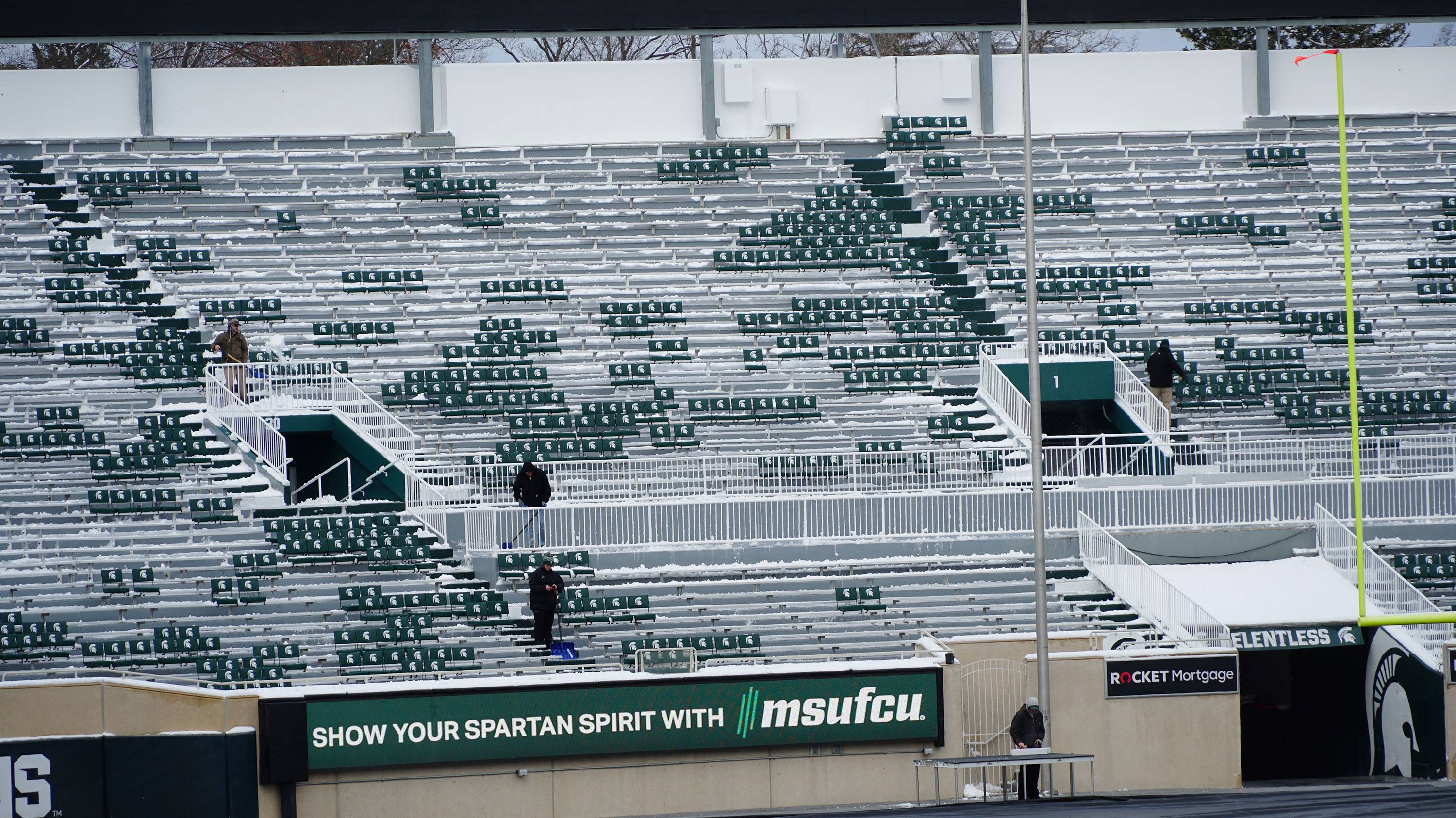A photograph shows several workers with shovels clearing the snow off the empty stands of Spartan Stadium.