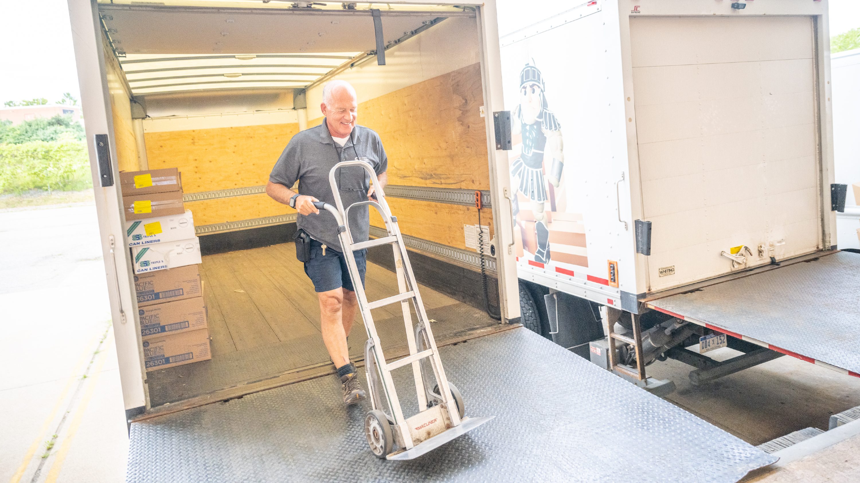 A man in a gray t-shirt and shorts pushes a hand truck out of the back of a cargo truck.