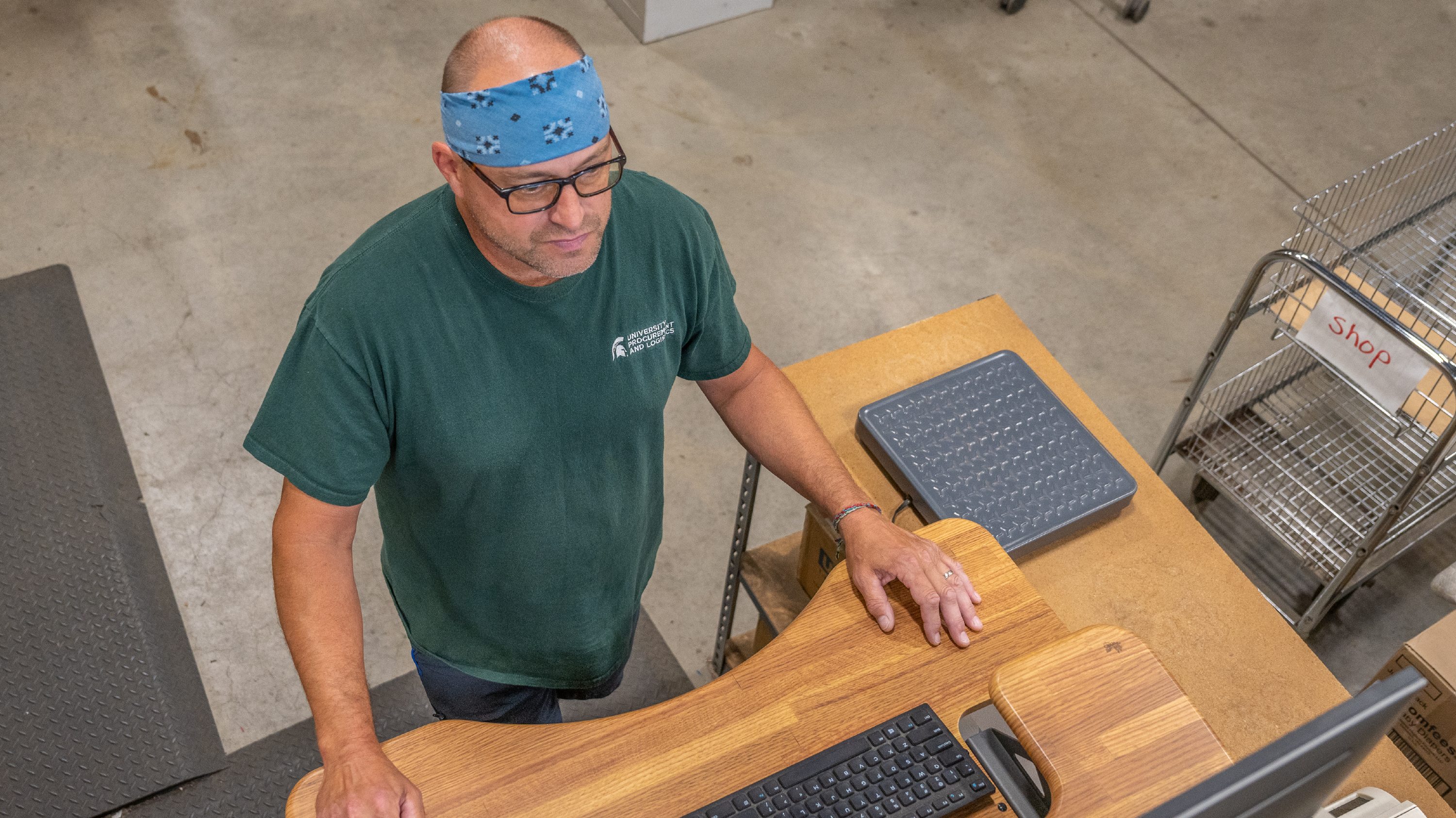 Viewed from above, a man in a green T-shirt and blue bandana operates a computer.